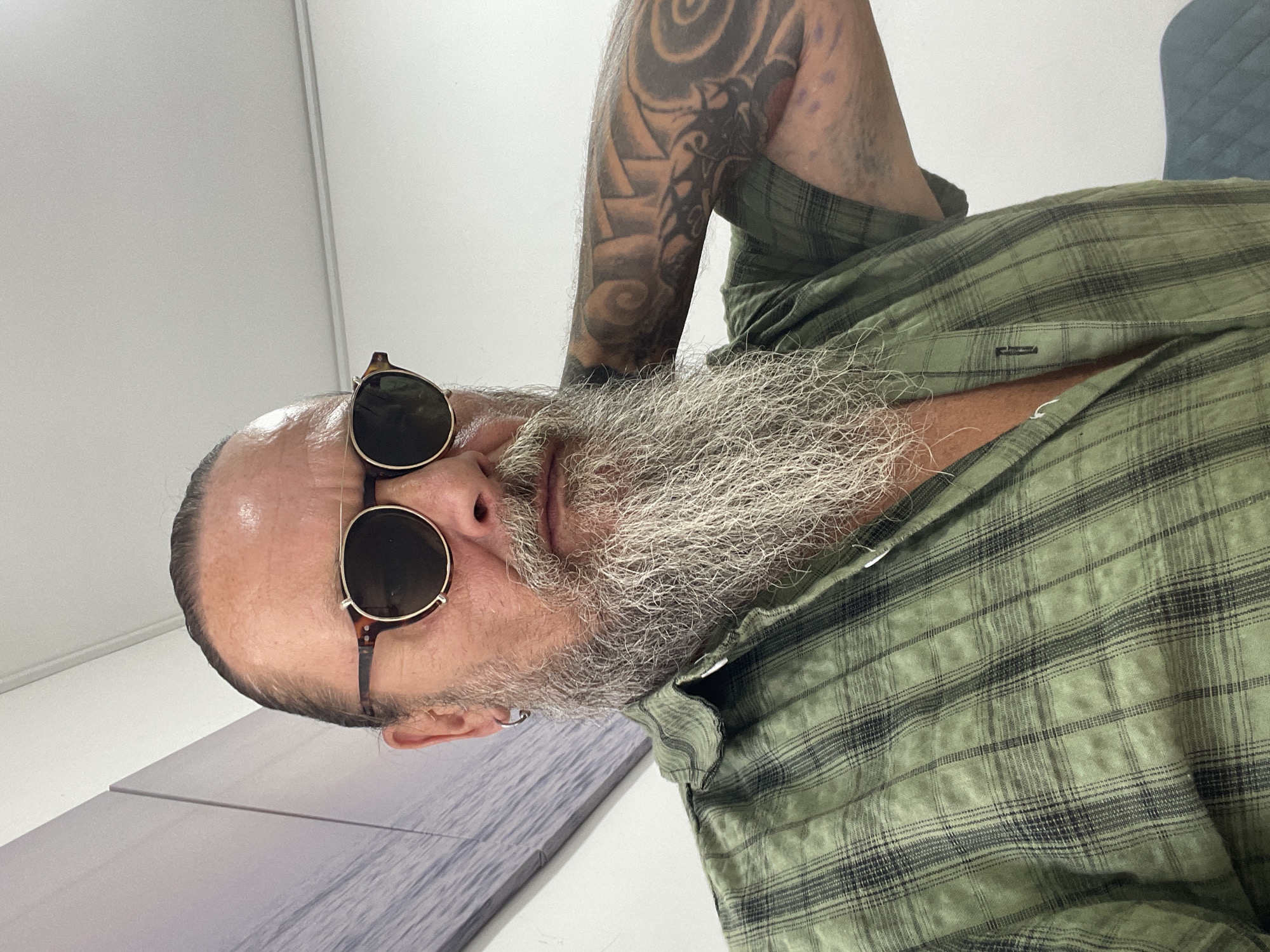 A man with a long, grey beard and sunglasses is looking at the camera. He is wearing a green plaid shirt with the top buttons undone, revealing his chest. He has tattoos on his arm, and he is holding his neck with his arm. The background is a white wall with a large artwork on the left side of the image. The artwork appears to be a depiction of a body of water with a light sky. The man's left ear has a silver hoop earring. The setting appears to be indoors, in a room that might be in Netanya, Israel.