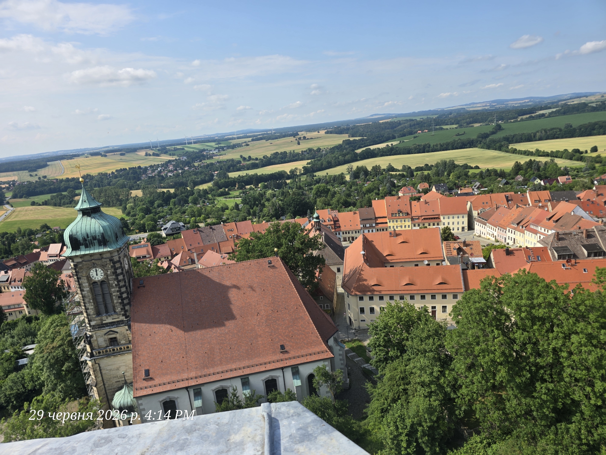 This media file is a photograph taken from an elevated viewpoint, offering a panoramic view of a town nestled within a rural landscape. The foreground is dominated by the spire and clock tower of a stone church, distinguished by its green copper roof. Adjacent to the church is a large building with a red tiled roof. The town itself features numerous buildings with terracotta-colored roofs, indicative of European architecture. Beyond the town, rolling hills stretch into the distance, interspersed with green fields and trees. Several wind turbines are visible on the horizon, suggesting a modern element within the landscape.
The scene depicts a tranquil daytime setting. The sky is a bright blue with scattered white clouds, indicating fair weather. The lighting suggests it is either late morning or early afternoon. The overall impression is one of a picturesque and serene location, likely a historic German town.
Visible text in the lower left corner reads "29 червня 2026 р. 4:14PM", which is Ukrainian for "June 29, 2026, 4:14 PM". This text appears to be a timestamp or watermark overlaid on the image. There is no discernible activity or event taking place; it is a static landscape photograph.