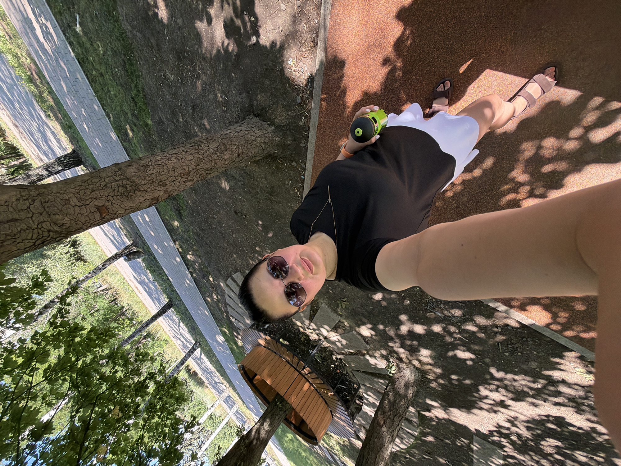 A woman in a black dress and white skirt is taking a selfie outdoors. She is wearing sunglasses and holding a green water bottle. Her feet are adorned with brown sandals. The woman is standing on a textured ground, possibly a park or outdoor recreational area, with dappled sunlight filtering through the trees. A path and grassy area are visible in the background, suggesting a natural or landscaped environment. The overall mood seems relaxed and casual, indicative of a sunny day. The time of day appears to be daytime, given the strong shadows. No visible text is present in the image.