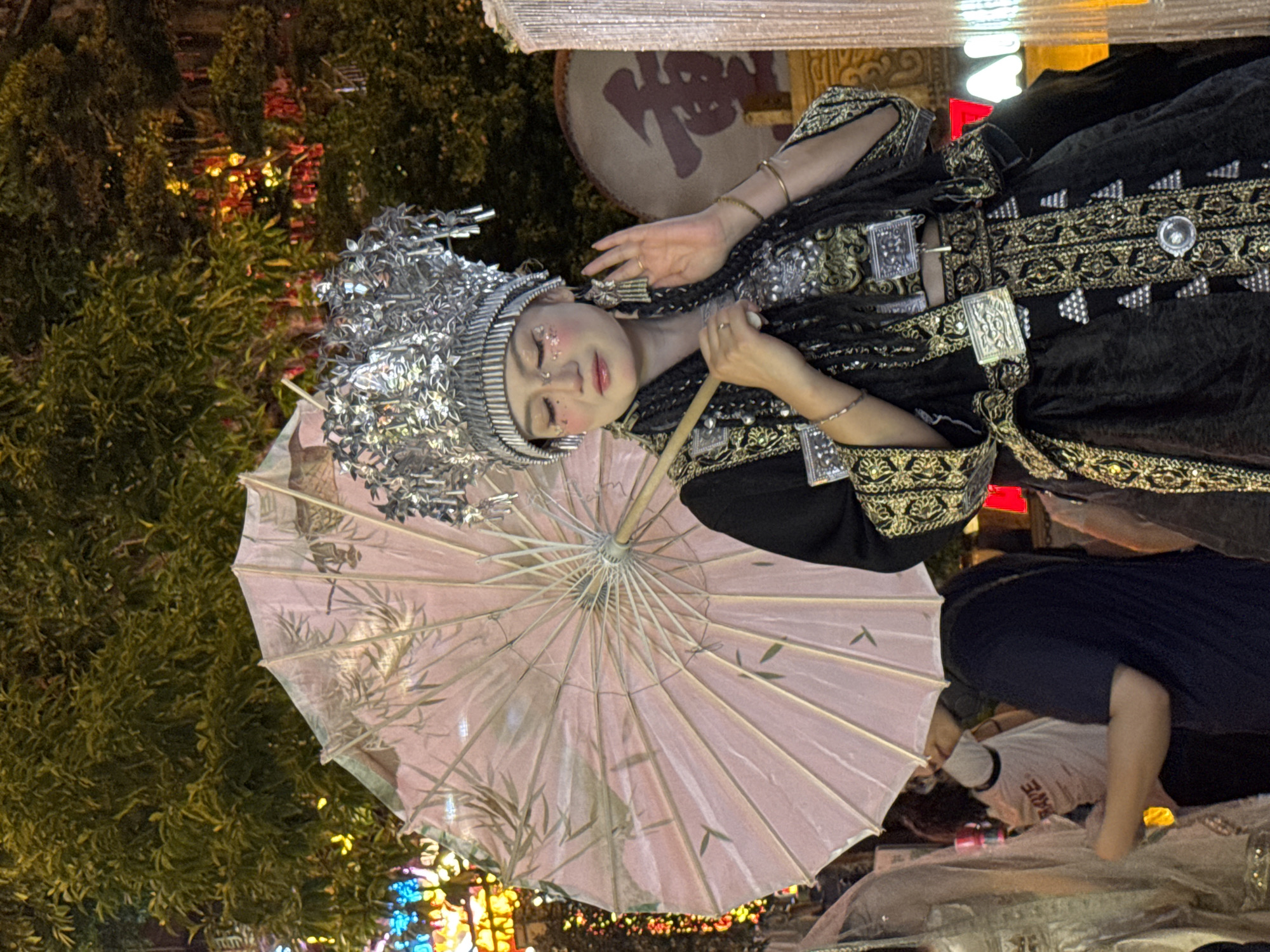 In Yuzhong District, China, a woman adorned in traditional festive attire stands holding a pink umbrella. Her headdress is elaborate and silver, and her clothing is black with gold and silver embellishments. She wears eye makeup with glitter. She is posed with her right hand raised, fingers splayed, and her left hand holds the umbrella's handle. Behind her, trees are visible, illuminated by colorful lights. To her right, a large drum with a Chinese character is partly visible. Other individuals and decorations are present in the background, suggesting a festive outdoor event.