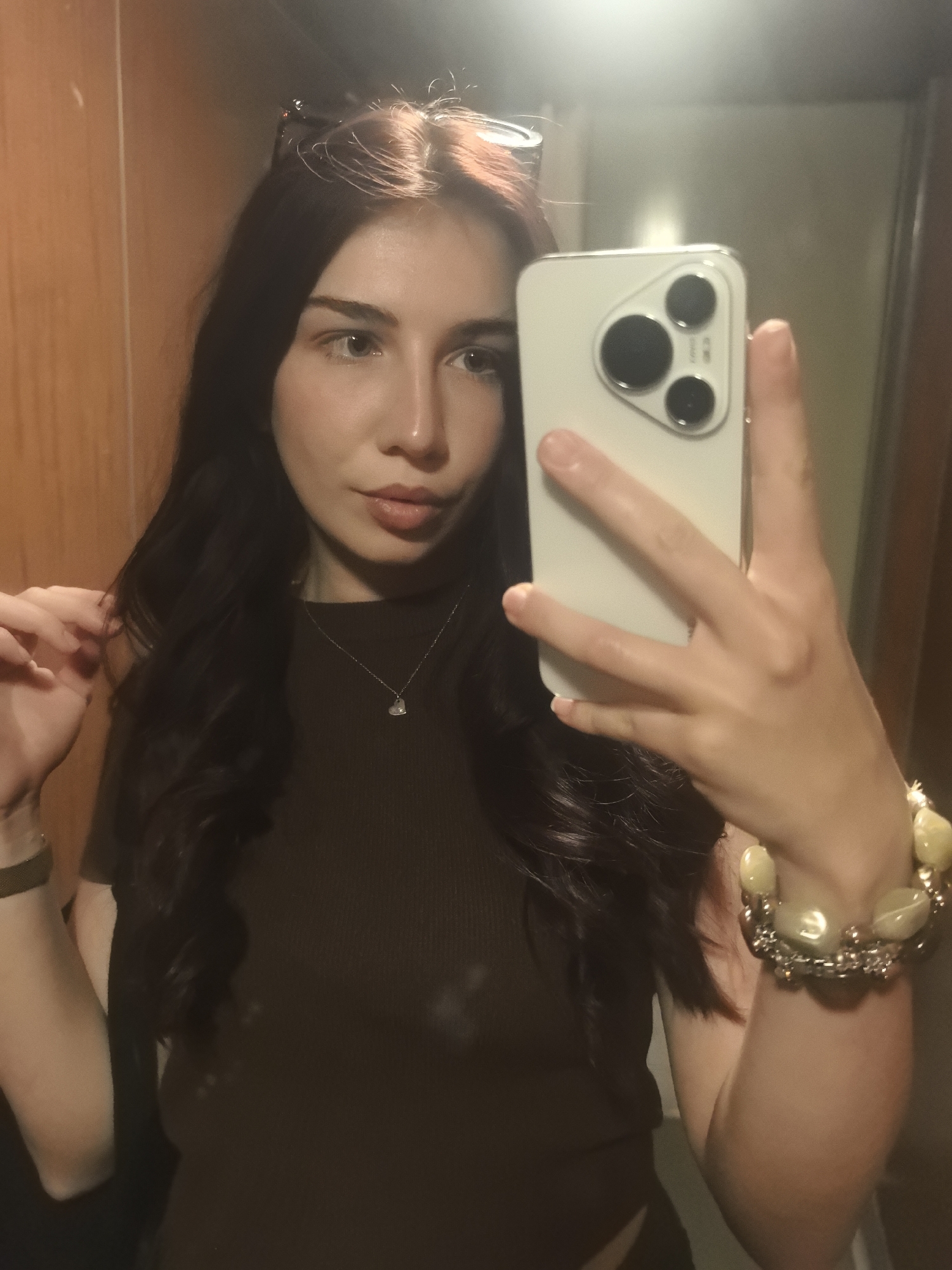 A woman is taking a selfie indoors, presumably in a private setting such as a home or a hotel room, given the presence of wood paneling. She is holding a white smartphone with a multi-lens camera system, which is obscuring part of her face. Her long, dark hair is styled in loose waves. She is wearing a dark brown top and a delicate necklace with a pendant. On her right wrist, she sports a beaded bracelet. Sunglasses are perched atop her head. The location of the scene is in the city of Unknown, Unknown.