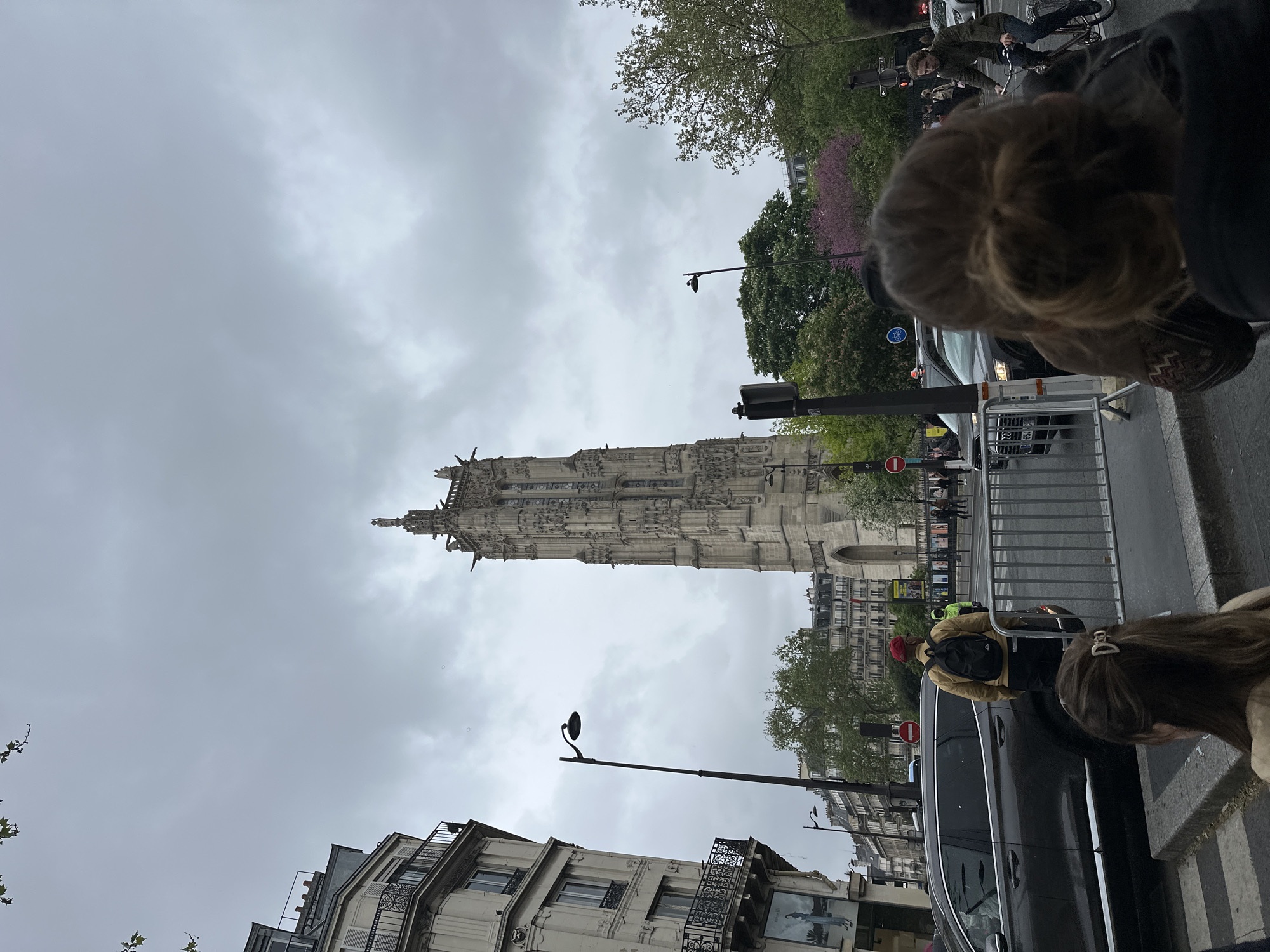 Here is a description of the image:
The image shows the Tour Saint-Jacques in Paris, France on a cloudy day. The tower is the focal point, a tall and ornate structure. Street-level views include Parisian buildings, street lamps, traffic lights, part of a street with a black car and people walking. There are trees with budding green leaves visible in the background, as well as a hint of pink blossoms on a tree. In the foreground, there is a metal security barrier, and portions of people’s heads/hair are in the foreground, potentially capturing the scene from an observer's perspective. Cyclists and pedestrian are also visible.