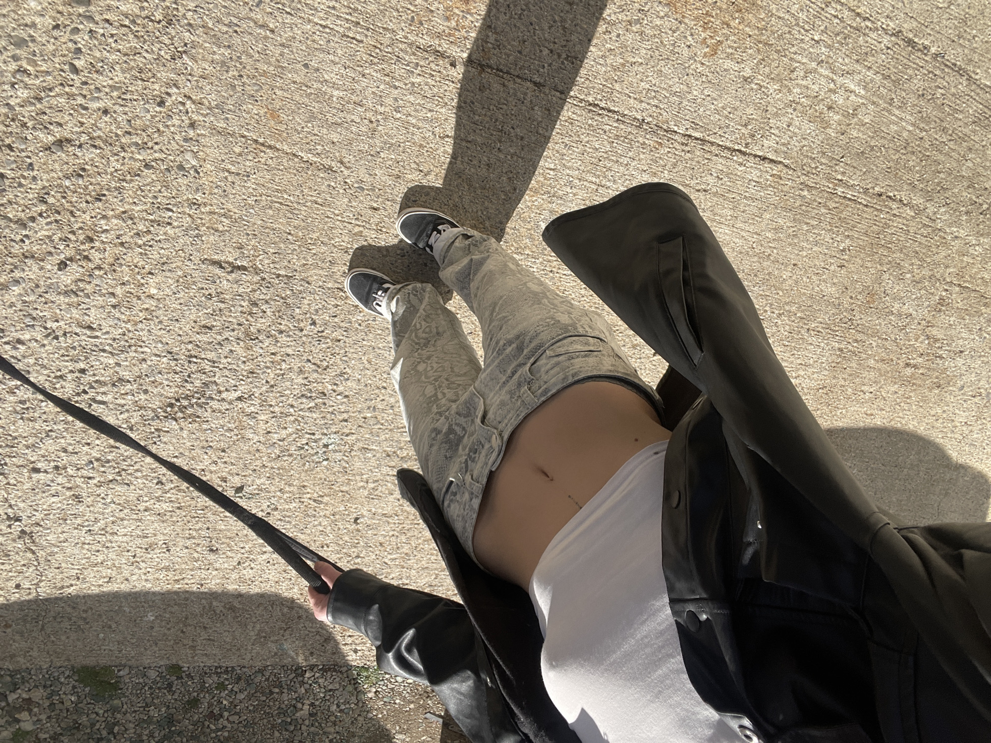 An overhead view captures a person standing on a light-colored, aggregate-exposed concrete surface in Boreti, Montenegro, under direct sunlight. The individual wears a black, open leather jacket over a white cropped tank top, exposing the midriff which shows a small mark or piercing. They are also wearing light grey patterned jeans and black low-top sneakers with white laces. The person's right hand holds a black strap or leash extending out of frame to the right. A distinct shadow of the person is cast forward onto the concrete, indicating the sun's position. In the upper right background, a rougher ground area composed of small pebbles and sparse green vegetation is visible.