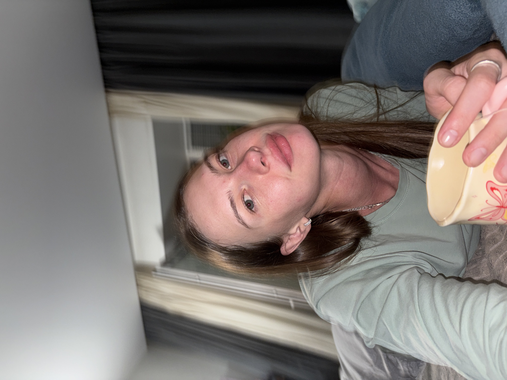 A woman is lying in bed and looking at the camera. She has long brown hair and is wearing a light green long-sleeved shirt. She is holding a mug with both hands. The room appears to be a bedroom with curtains in the background. The location is not specified as Perm, Russia in the image.