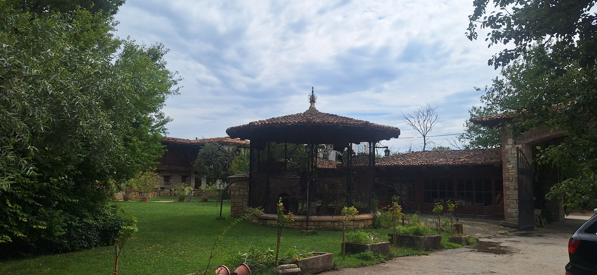 The image depicts a courtyard area with a traditional Bulgarian architectural style, possibly located in or near Kalimantsi, Bulgaria. In the foreground, a circular, open-air pavilion with a tiled, octagonal roof stands prominently. The pavilion is enclosed by decorative metal fencing and has a stone base. Surrounding the pavilion is a well-maintained green lawn with several potted plants and stone planters containing vegetation.
In the background, several buildings with red tile roofs and stone and wood facades are visible. These structures appear to be part of a historical complex or estate. Trees and dense foliage flank the scene, suggesting a rural or semi-rural setting. The sky is partly cloudy, with white and grey clouds against a pale blue background. The lighting suggests it is daytime. There are no people or animals clearly visible in the image. A black car is partially visible on the right side of the frame, indicating the presence of modern elements within this traditional setting. The overall impression is one of a preserved historical site or a rustic resort.