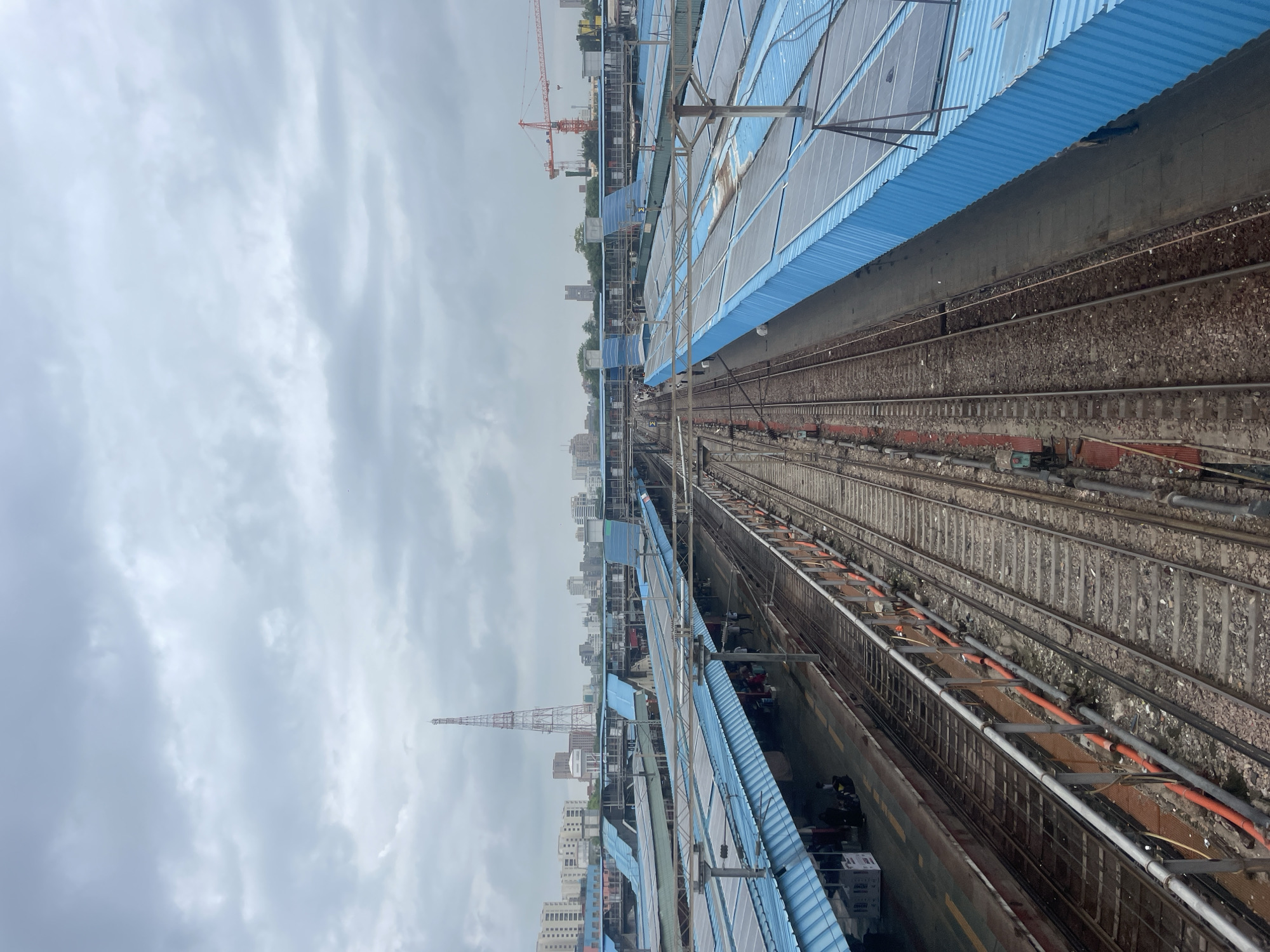 This is a high-angle, full shot of a busy train station in New Delhi, India, under a cloudy sky. The scene is dominated by multiple train tracks and platforms covered by bright blue canopies. Many of these canopies are equipped with solar panels, reflecting the sky. The platforms show scattered groups of people, some with luggage, suggesting a moderate level of activity. Visible structures include a tall radio tower in the background, several buildings of varying heights, and an orange construction crane. The train tracks are well-maintained, with ballast and sleepers clearly visible, and some are lined with railings and conduits carrying cables. The overall atmosphere is utilitarian and functional, typical of a major urban transportation hub.