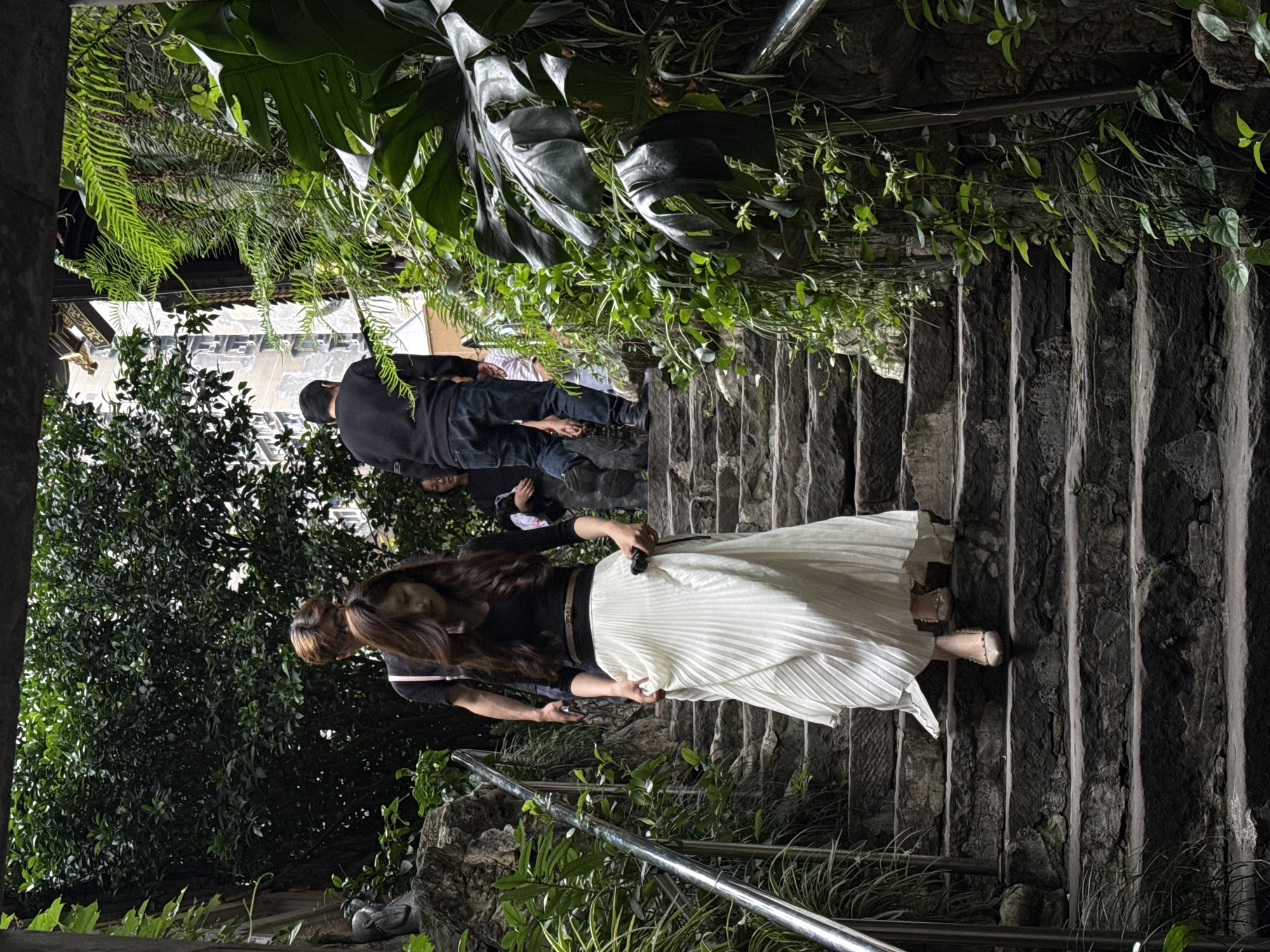 A medium, slightly rotated, full outdoor shot depicts a woman in a white pleated skirt walking down a set of stone stairs in Yuzhong District, China. The steps are steep, and a chrome handrail is visible on the lower left. The woman is wearing a black top, and the wind slightly billows her skirt. A man in a black shirt is standing behind her. Lush greenery surrounds the stairs, with ferns and broadleaf plants above and dense foliage to the left. The scene appears to be a blend of urban architecture and natural elements.