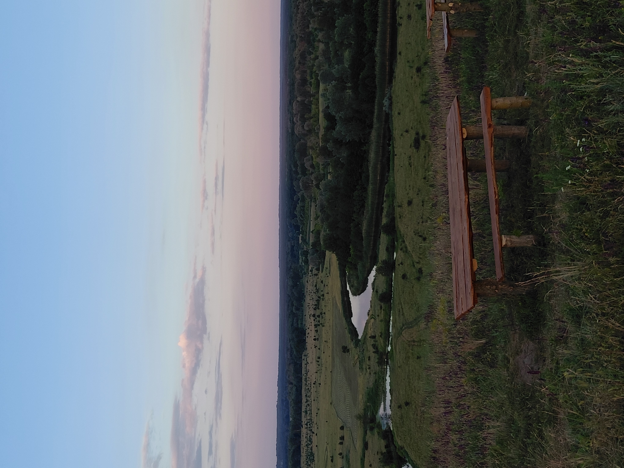 The image shows a landscape view of Ustyvytsia, Ukraine at dusk. In the foreground, there is a wooden picnic table and bench set on a grassy knoll. Beyond the knoll, a winding river is visible, flowing through a valley dotted with trees. The background consists of a dense forest that stretches to the horizon, under a pale blue and pink sky with scattered clouds. The scene is serene and natural.