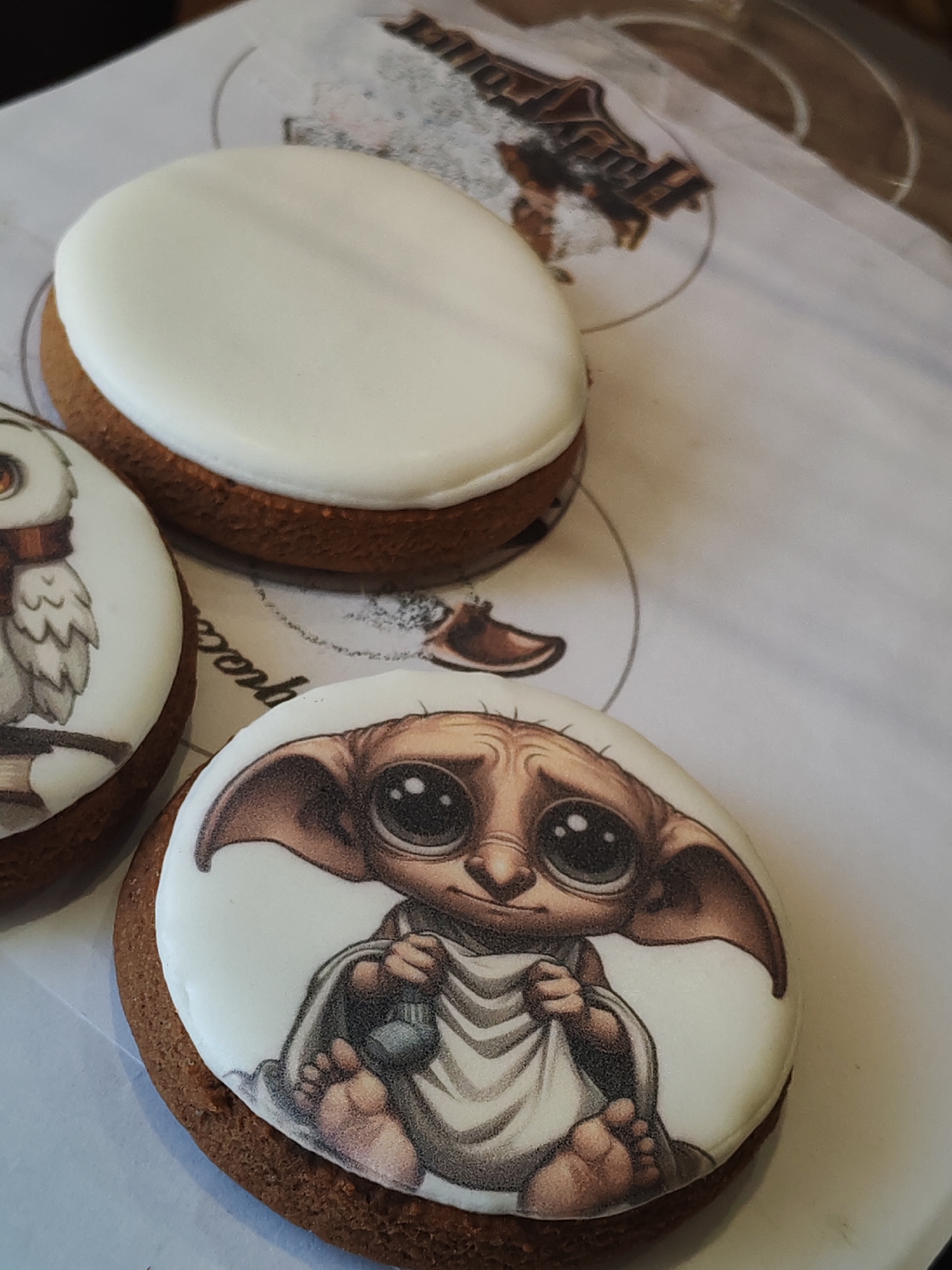 Three round cookies are arranged on a white surface printed with Harry Potter-themed imagery. The top cookie is plain white, while the bottom two have images printed on them. One cookie features an owl, and the other depicts Dobby the House Elf. The cookies are on display in Unknown, Unknown.