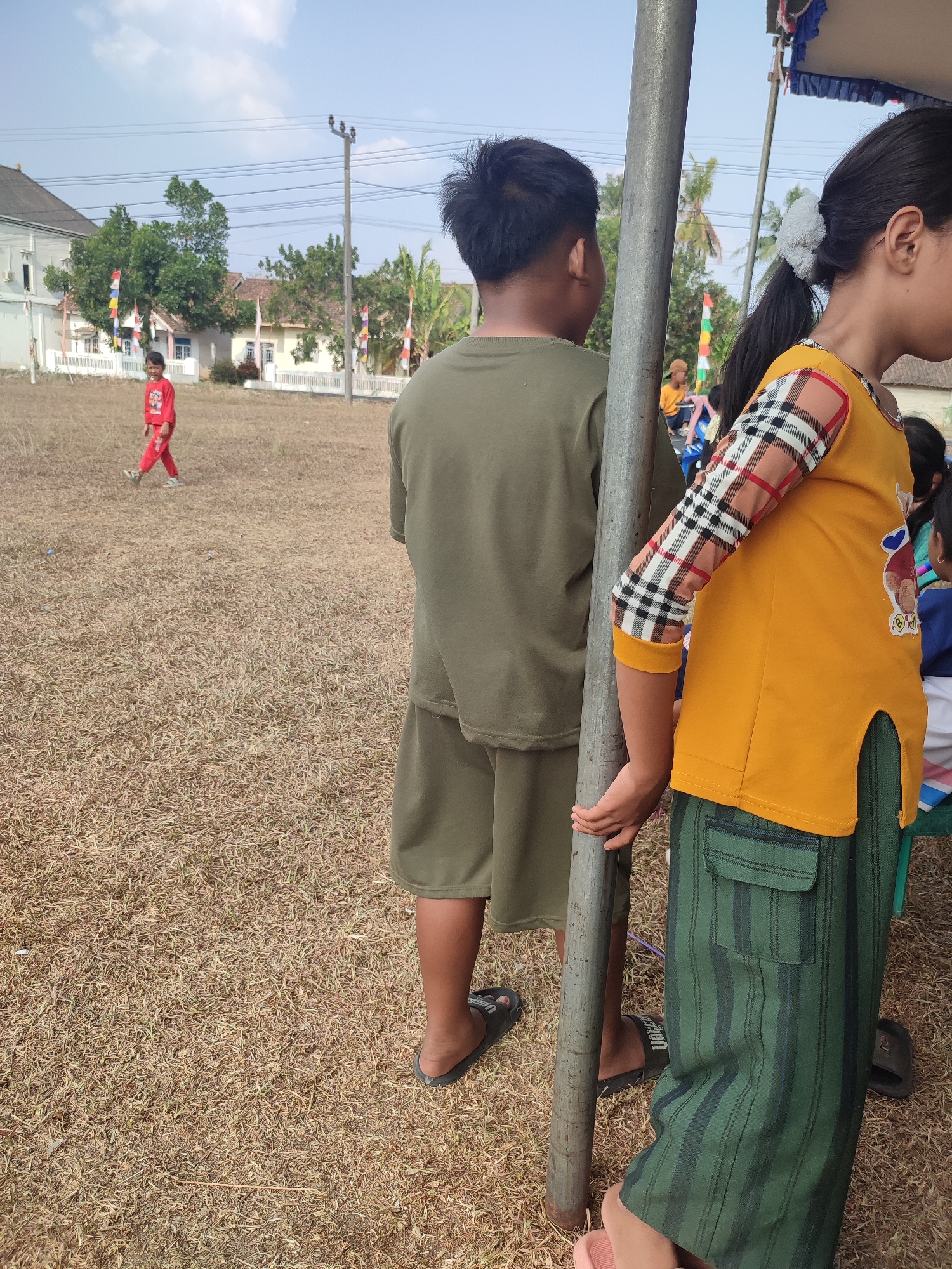 In Nusa Jaya, Indonesia, an outdoor scene unfolds on a dry, brown grassy field under a bright, partly cloudy sky. Two children are the central focus. A boy, facing away from the camera, wears a khaki-colored t-shirt and shorts. Beside him, a girl, also facing away, sports a yellow long-sleeved shirt with a checkered pattern on the sleeves and green striped pants. She is holding onto a metal pole. In the background, a red-clad child walks across the field, and several houses with white fences and red-and-white Indonesian flags are visible. Trees and palm trees dot the landscape, and electrical poles with wires are present. The overall impression is a casual gathering or event in a rural or suburban Indonesian setting.
