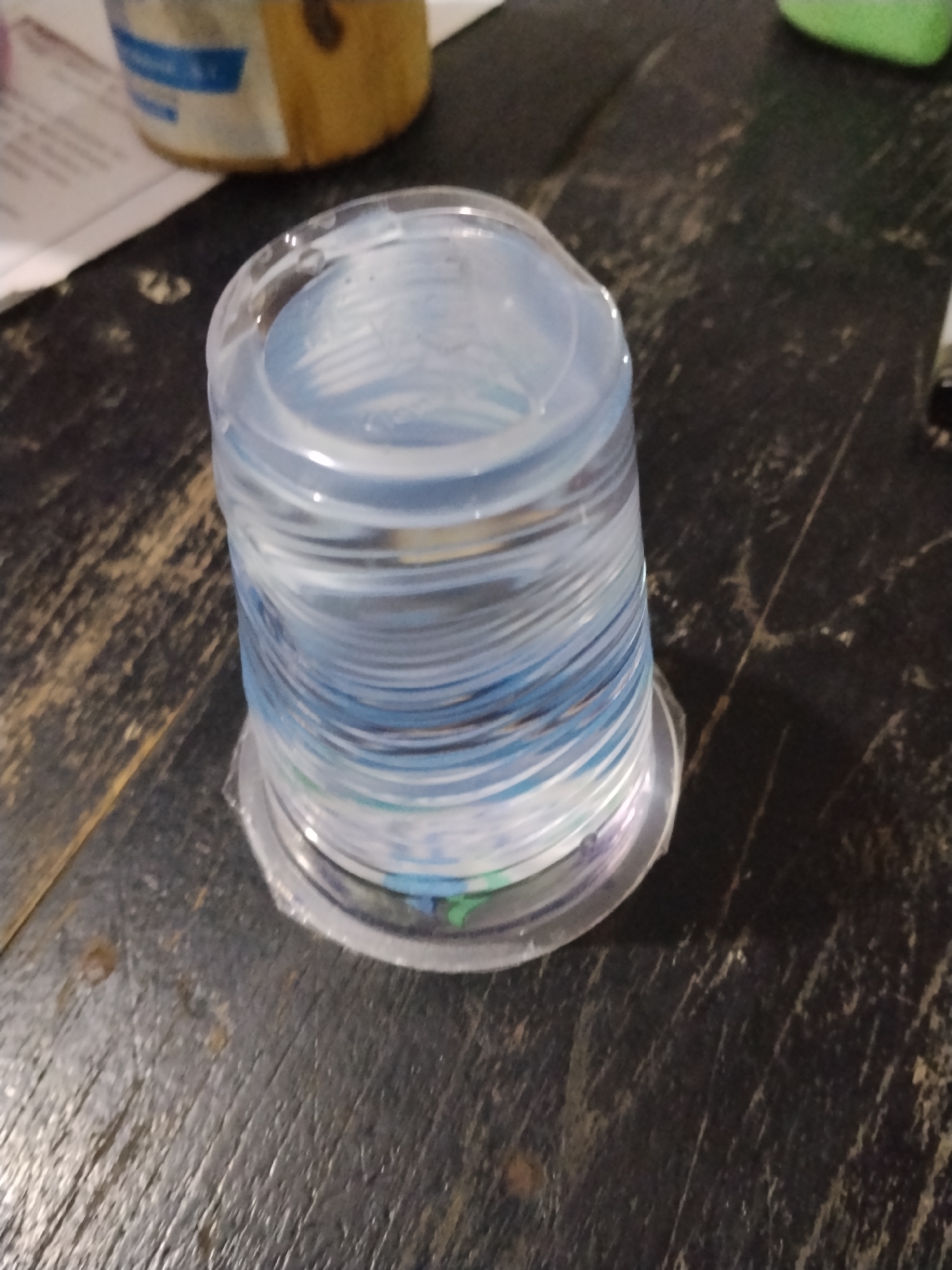 The image shows a stack of clear plastic cups filled with a light blue liquid, likely water. The cups are stacked in a cylindrical form and are sitting on a dark, textured surface, possibly a wooden table with a worn finish. The lighting is dim, casting shadows and highlighting the ripples in the liquid and the texture of the surface. There are some other out-of-focus objects in the background, including a partially visible container with a blue label and a blurry green object. The overall impression is a casual, indoor setting. No specific location within Bilis Bilis, Indonesia, can be determined from the image alone.