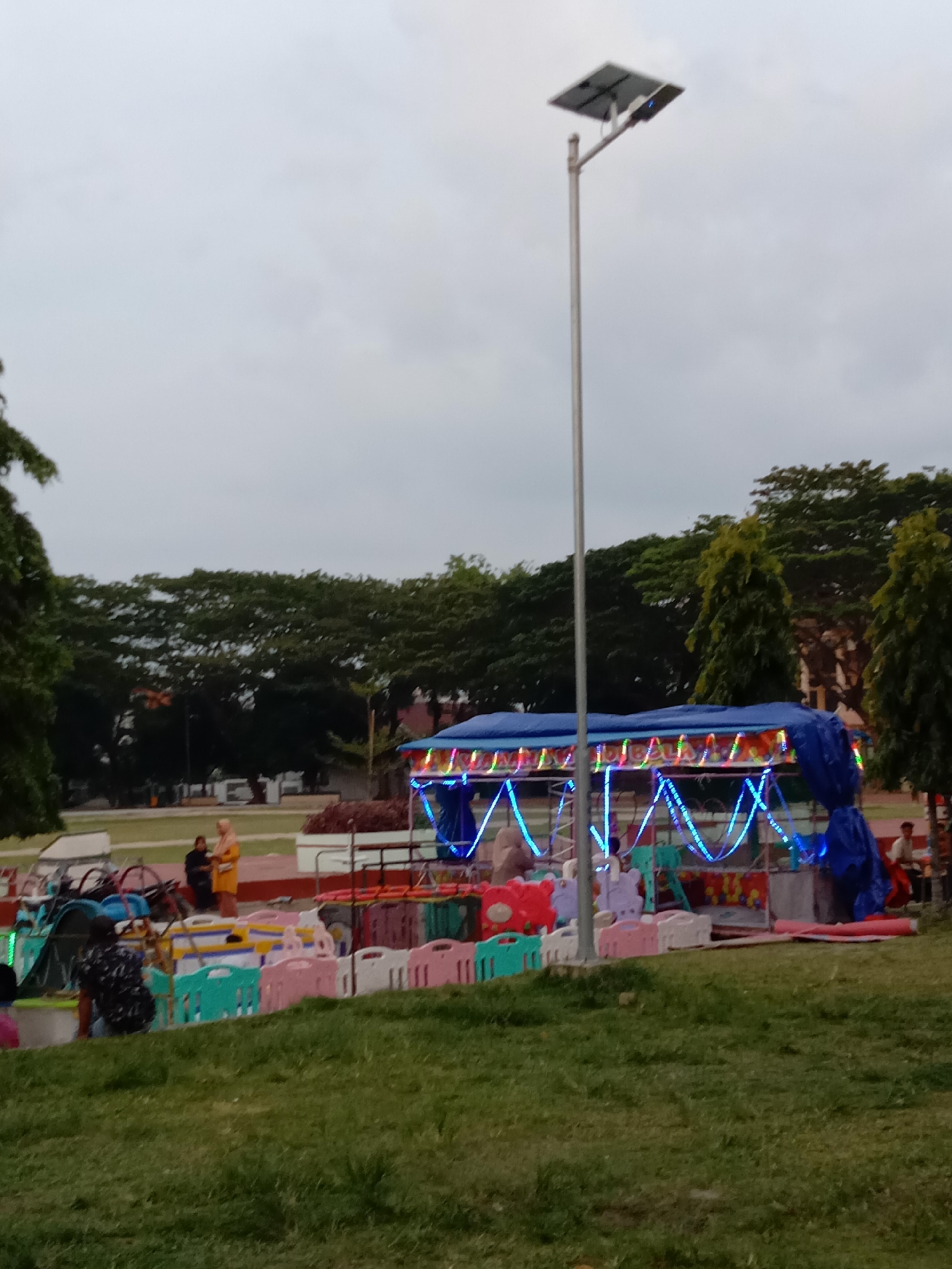 This image captures an outdoor scene, likely a public park or recreational ground, in Lhokseumawe, Indonesia. The sky is overcast, indicating a cloudy day, late afternoon, or early evening.
In the mid-ground, a central blue-tarped structure with vibrant string lights (blue, green, red, yellow) suggests a temporary stall or children's amusement ride. In front of this, low, colorful plastic barriers in shades of pink, white, and light green create an enclosure. Behind these barriers, several small pedal-powered vehicles, such as tricycles or bicycles, are visible, reinforcing the idea of children's rides.
Several people are present in the scene. In the mid-ground, two women stand near the rides; one is dressed in a yellow outfit and headscarf, while the other wears darker clothing. Another person in a patterned shirt is partially visible in the left foreground. To the far right, a person is seated on a red bench-like structure.
A tall, silver street light pole with a solar panel mounted on top stands prominently in the center-right of the frame, reaching towards the cloudy sky. The background is filled with a dense line of green trees, providing a natural backdrop, with some taller, slender trees also scattered. Faint outlines of distant buildings are visible through the trees. The foreground consists of a large expanse of green grass. The overall scene suggests a casual community gathering or a small local fair. There is no visible text in the image.