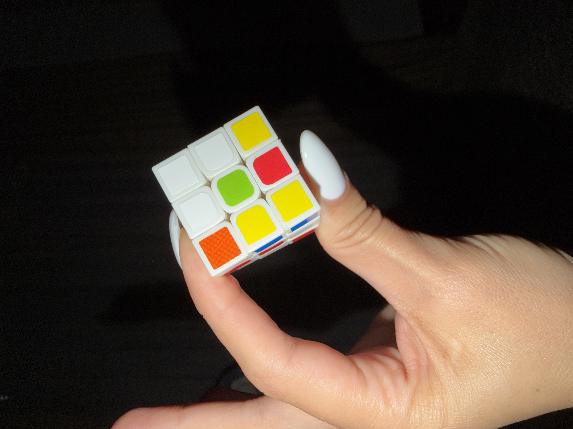 A hand with a white fingernail is holding a partially solved Rubik's Cube in a dark environment. The cube has white, green, yellow, red, and orange squares visible. The background is dark and out of focus. There is no information provided in the image to determine if this scene is located in Budva, Montenegro.