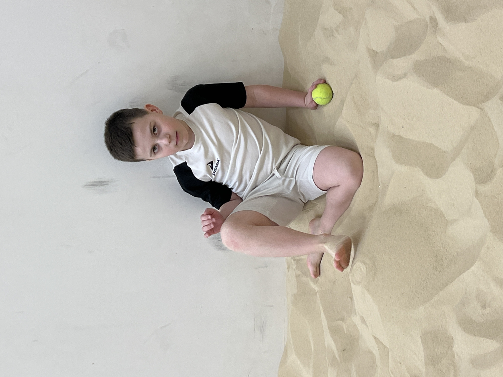 A male child, approximately 8-12 years of age, with short brown hair, is seated on an indoor artificial sand court. He maintains direct eye contact with the viewer, exhibiting a neutral facial expression. He is wearing a short-sleeved athletic shirt, white with black raglan sleeves, featuring a "DEMIX" logo on the left chest, and light-colored shorts. He is barefoot. His left leg is bent with the knee elevated, while his right leg is bent laterally. His right arm is extended downward, with his hand resting on the sand and loosely holding a single fluorescent yellow tennis ball. The background consists of a plain, light-colored (off-white/light grey) wall displaying minor scuff marks. The environment is an indoor sports facility with a deep layer of light beige sand forming dunes, characteristic of an indoor beach sports arena. This scene is located within a specialized indoor beach sports facility in Saint Petersburg, Russia, consistent with venues such as Volleygrad or the Arena Saint Petersburg Beach Sports Center.