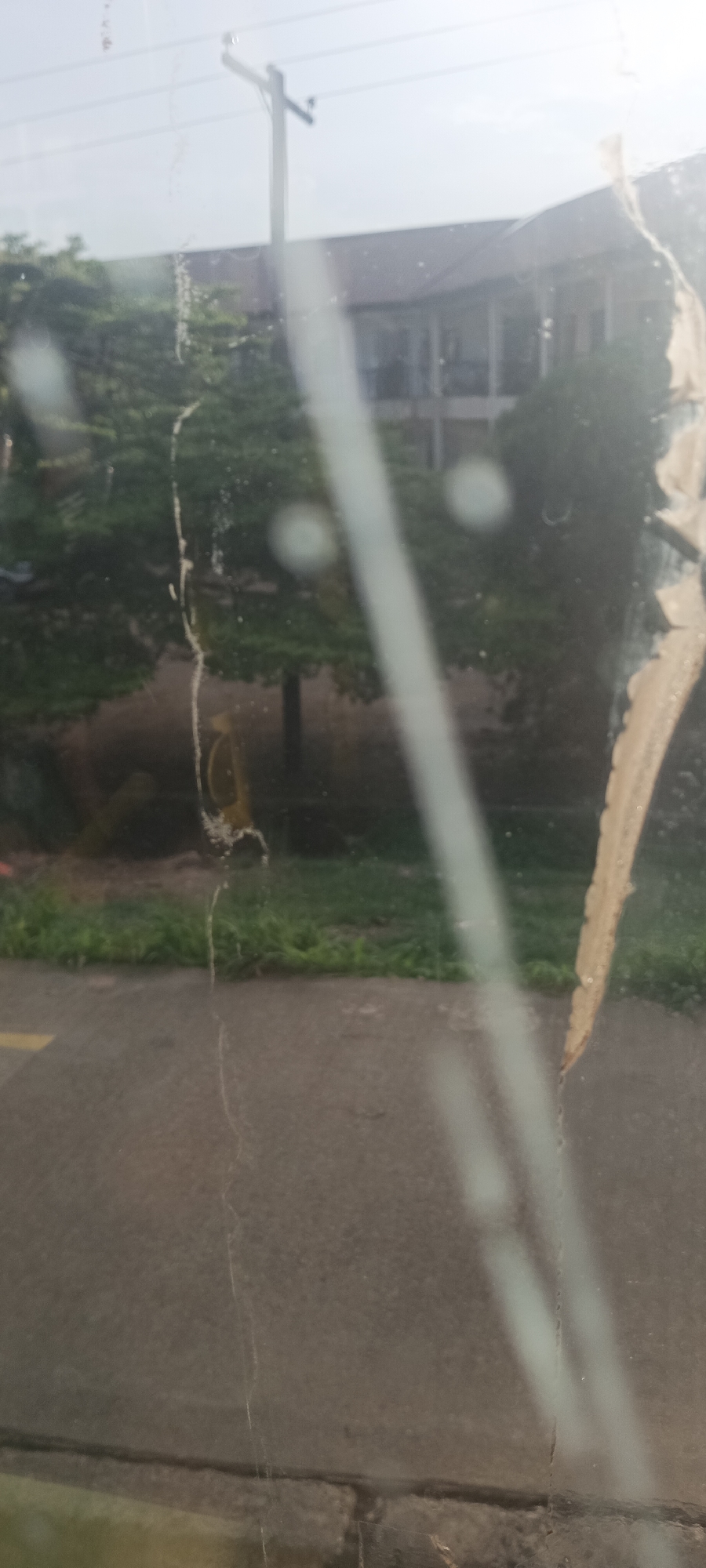 The image presents an outdoor scene viewed through a heavily smudged, scratched, and dirty window or glass pane. The foreground is dominated by prominent vertical and irregular streaks, as well as patches of residue on the glass. A long, light-colored, slightly curved streak runs vertically through the center, while a wider, irregularly shaped, light brown or beige smudge, possibly dried matter, is visible on the right. A thinner, textured vertical streak appears on the left. Two blurry, circular, bright spots are positioned in the upper-middle, resembling reflections or light sources.
Through the obscured glass, a daytime environment is visible. In the background, a multi-story building with a dark roof and lighter walls is partially discernible, interspersed with numerous green trees. A utility pole with crossarms and wires stands tall on the upper left side. In the lower half, a grey asphalt or concrete road stretches across the frame, bordered by a strip of green grass or weeds. Faint markings, possibly faded road lines, are barely visible on the road surface.
No specific activity or event is clearly taking place; the scene appears static. The bright, washed-out sky and lighting indicate it is daytime. There is no visible text in the image. The location context provided is Gura Top, Nigeria.