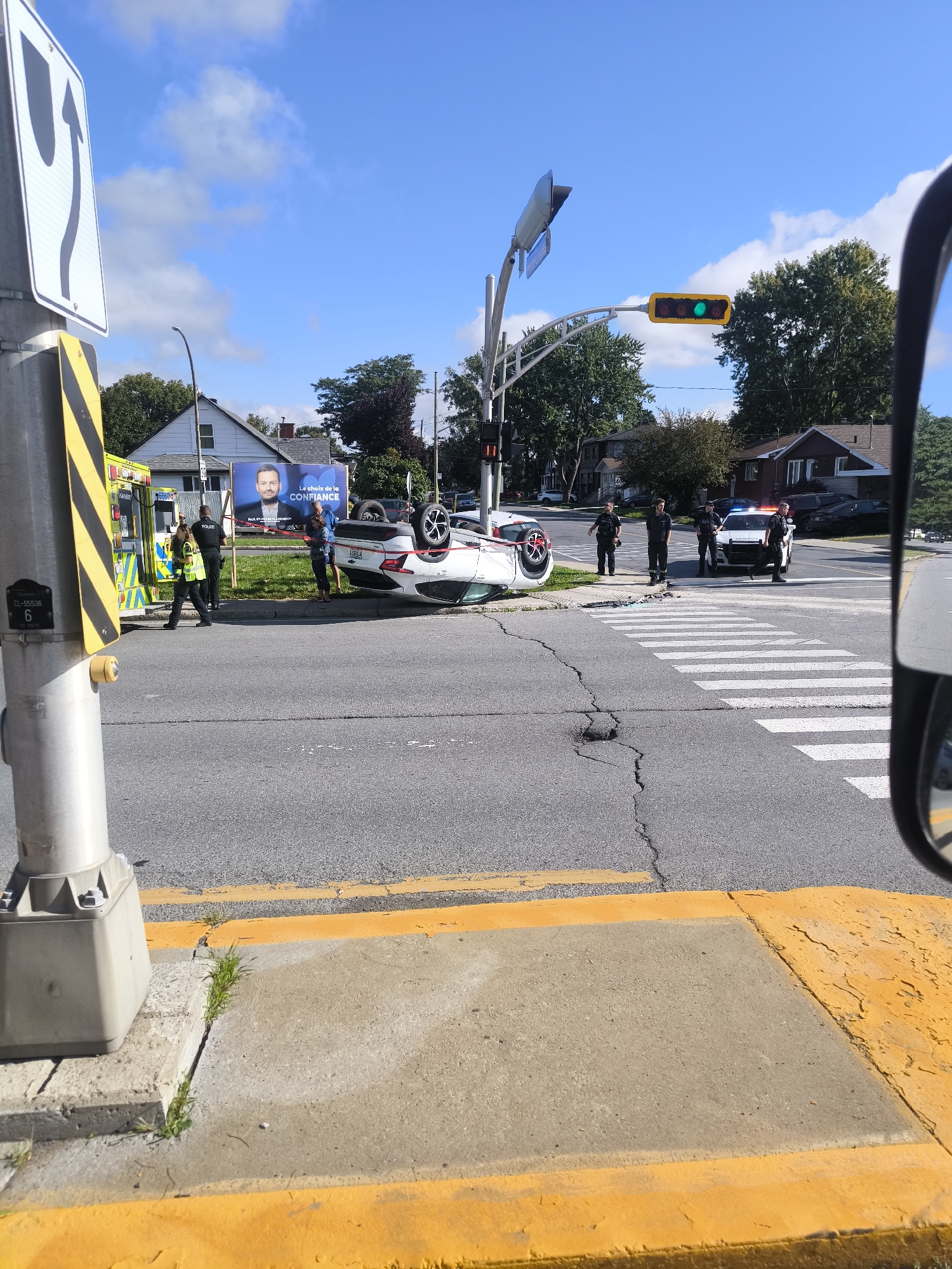 The image depicts the aftermath of a car accident at an intersection in Longueuil, Canada. A white car is overturned on its roof in the middle of the intersection, which has a pedestrian crosswalk. Several police officers are present, some standing near the overturned vehicle and others by a police cruiser parked nearby. An ambulance is also visible at the scene. Traffic lights are visible, with one showing a green light. A large billboard with a political candidate's image and campaign slogan is visible in the background. The scene appears to be on a sunny day, with a clear blue sky and some clouds. The environment includes residential buildings and trees along the street.
