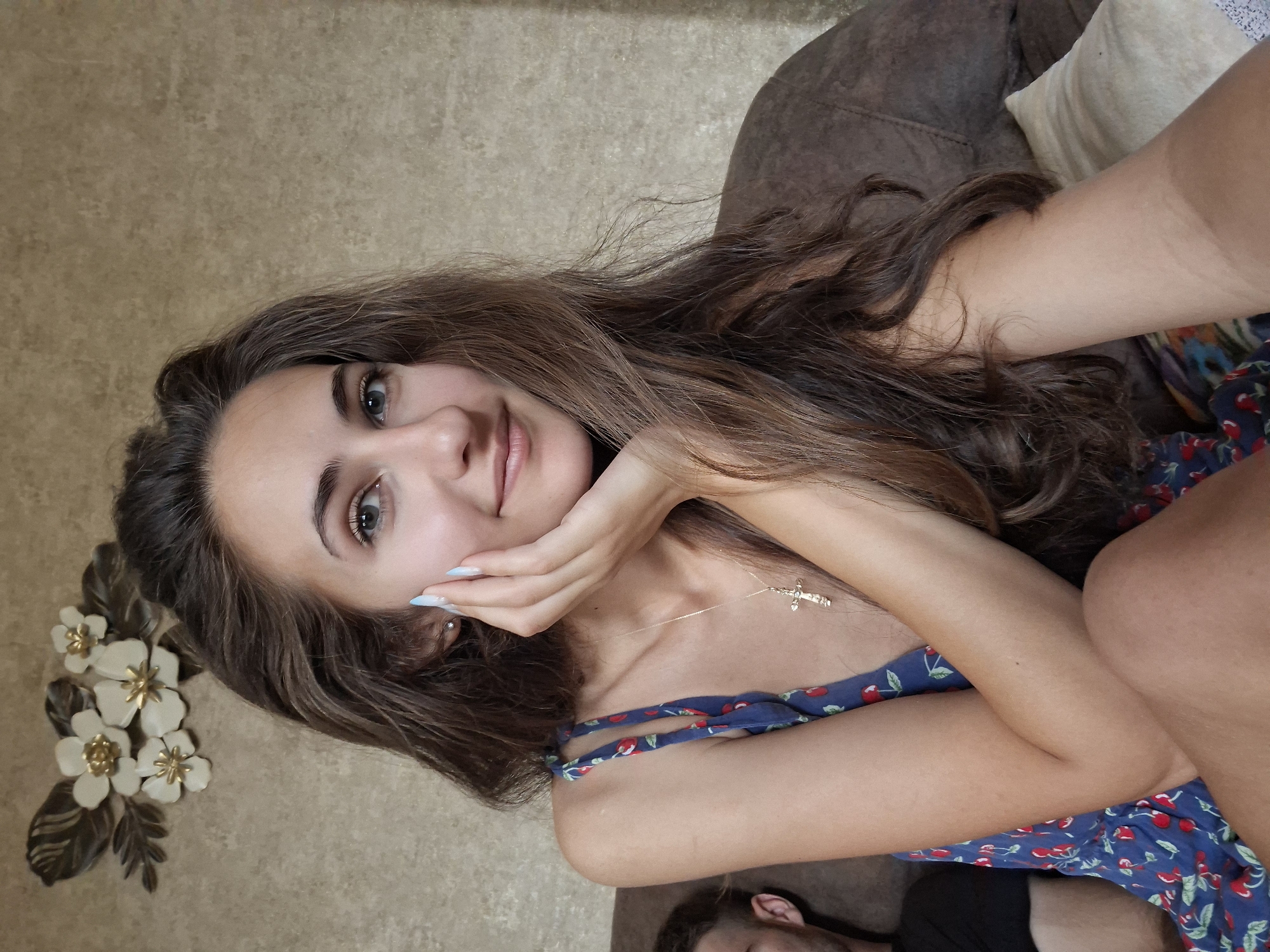 A young woman with long, brown, wavy hair is seated on a couch in an indoor setting. She is wearing a blue dress with a cherry pattern and a gold cross necklace. Her right hand is resting on her cheek. A man's left shoulder and head are visible to the left of the woman. Behind her, on a wall, is a floral wall ornament. The lighting is soft and natural.