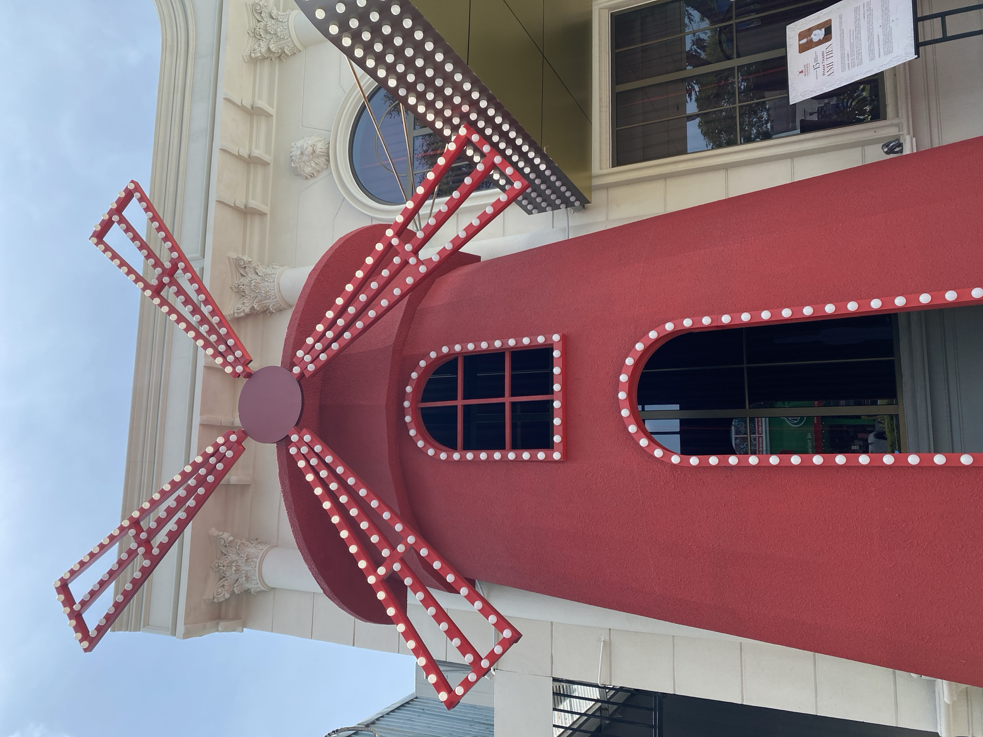 The image shows a red, windmill-shaped structure with lights on its sails, positioned in front of a white building with classical architectural elements. The building features columns, ornate carvings of lion heads, and arched windows. The windmill has an arched doorway and a square window, both outlined with lights. A sign is visible on an easel to the right of the windmill, with text in Vietnamese. The overall scene suggests a playful or artistic installation, possibly in a commercial or entertainment district. The location is identified as the Moulin Rouge complex in the city of Đà Nẵng, Vietnam, a modern architectural development that incorporates themed elements.
