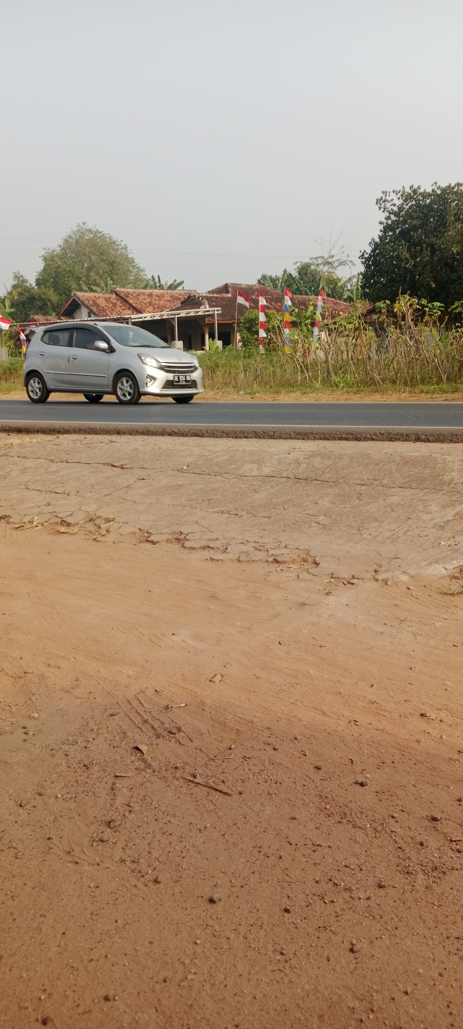 A silver hatchback car is driving on a paved road in Sidodadi, Indonesia. The car is positioned in the left lane, facing to the right of the frame. The road is bordered by a dirt embankment on the left and a grassy area with houses and trees on the right. Several Indonesian flags are visible in the background, suggesting a national holiday or celebration. The sky is overcast and the lighting is soft, indicating it might be morning or late afternoon. The foreground consists of a dry, dusty ground with some cracks and small stones.