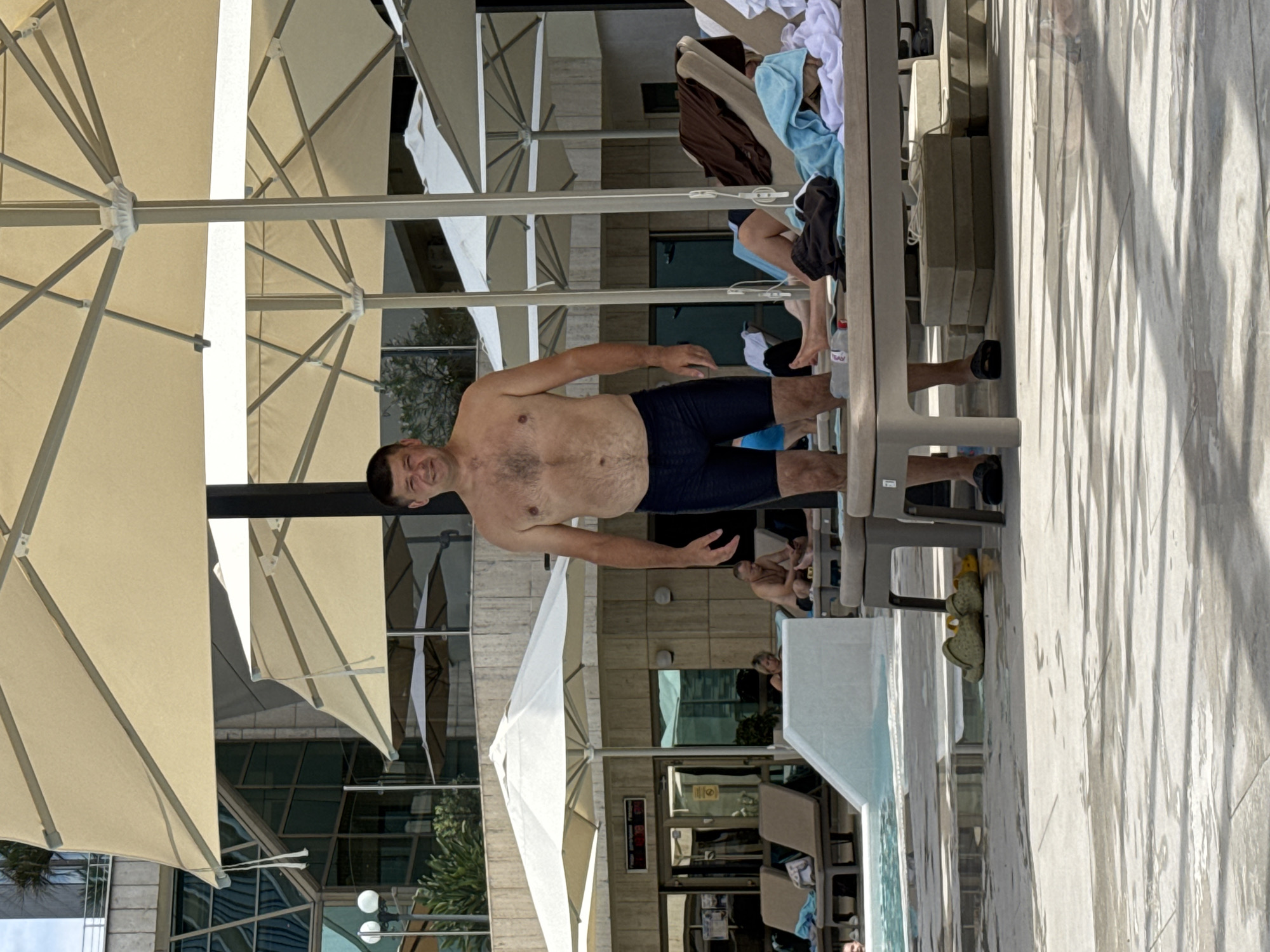 A man in dark blue swimming trunks stands on a wet tiled surface next to a swimming pool. He is looking at the camera and smiling. Behind him, several large beige umbrellas shade lounge chairs, some of which are occupied by other people. In the background, a modern building with a glass facade is visible. The scene appears to be a hotel or resort in Sochi, Russia, likely during a warm season.