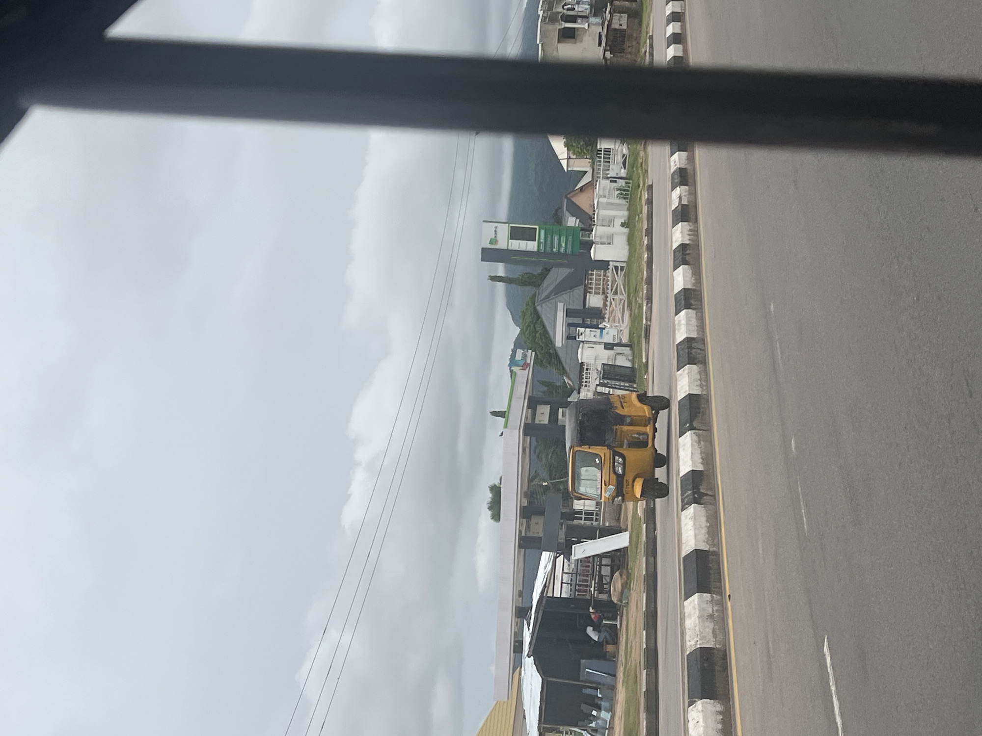 The image captures a view from a vehicle, looking out towards a street scene in Jalingo, Nigeria. In the foreground, a gray asphalt road is visible with a yellow and black striped concrete barrier separating it from the sidewalk. A yellow auto-rickshaw, known locally as a Keke Napep, is driving on the road. Beyond the barrier, there are several structures, including what appears to be a gas station with a white canopy and signage. To the left of the gas station, a small open-air stall or workshop is present, with people and materials visible inside. In the background, houses and trees are scattered, and a large, cloud-covered mountain range dominates the horizon. The sky is overcast with heavy clouds, suggesting a gray, possibly humid, day. The perspective is from inside a vehicle, indicated by the dark, out-of-focus bars framing the right side of the image.