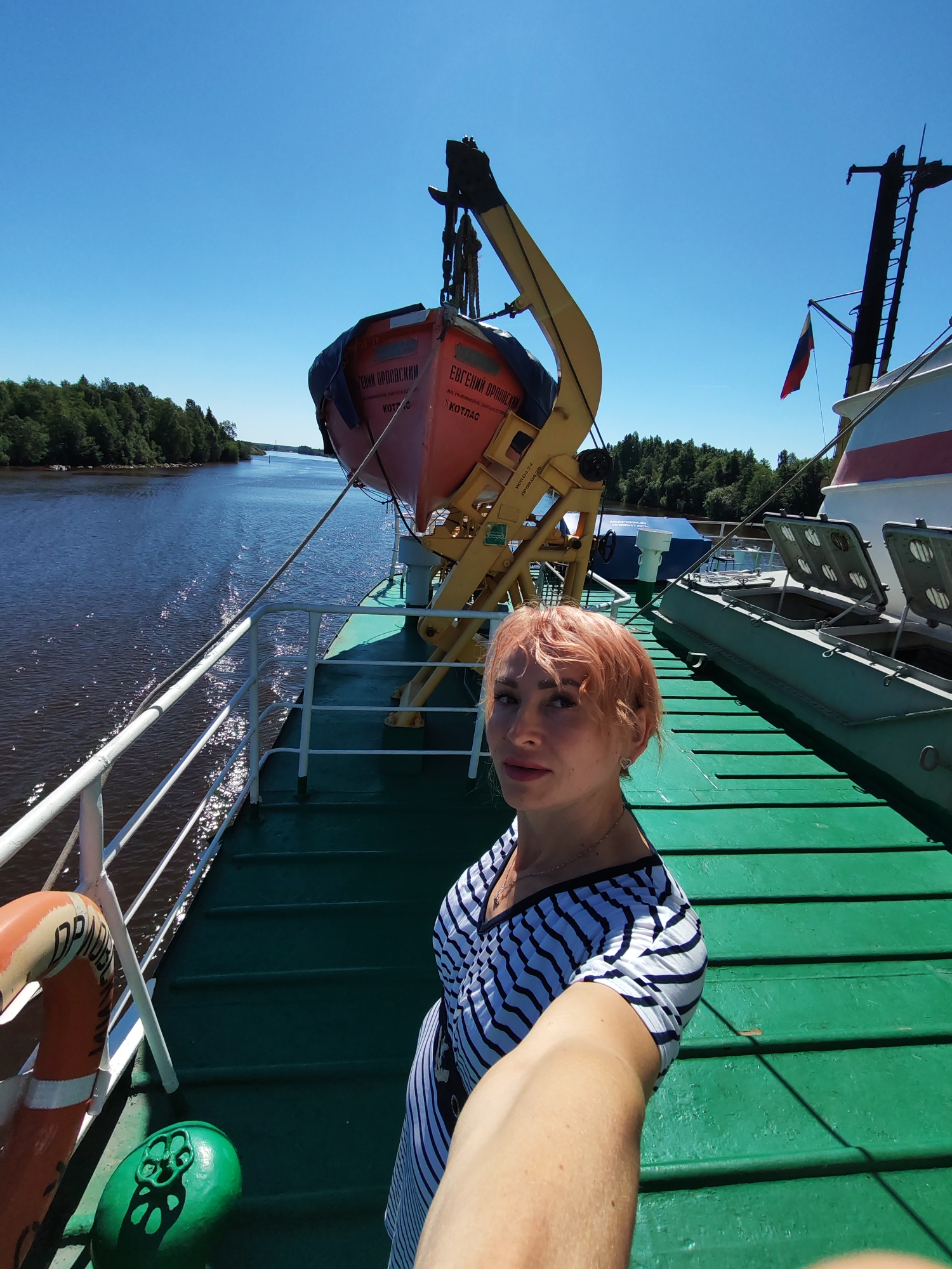 A woman with short, reddish-blonde hair is taking a selfie on the deck of a ship. She is wearing a black and white striped shirt. To her left, a life preserver is visible. In the background, a river is bordered by a dense forest, and the sky is clear blue. A lifeboat is suspended from a crane on the ship, with Russian text visible on it. A Russian flag flies from a mast on the ship. The deck of the ship is painted green. The image appears to have been taken on a sunny day.