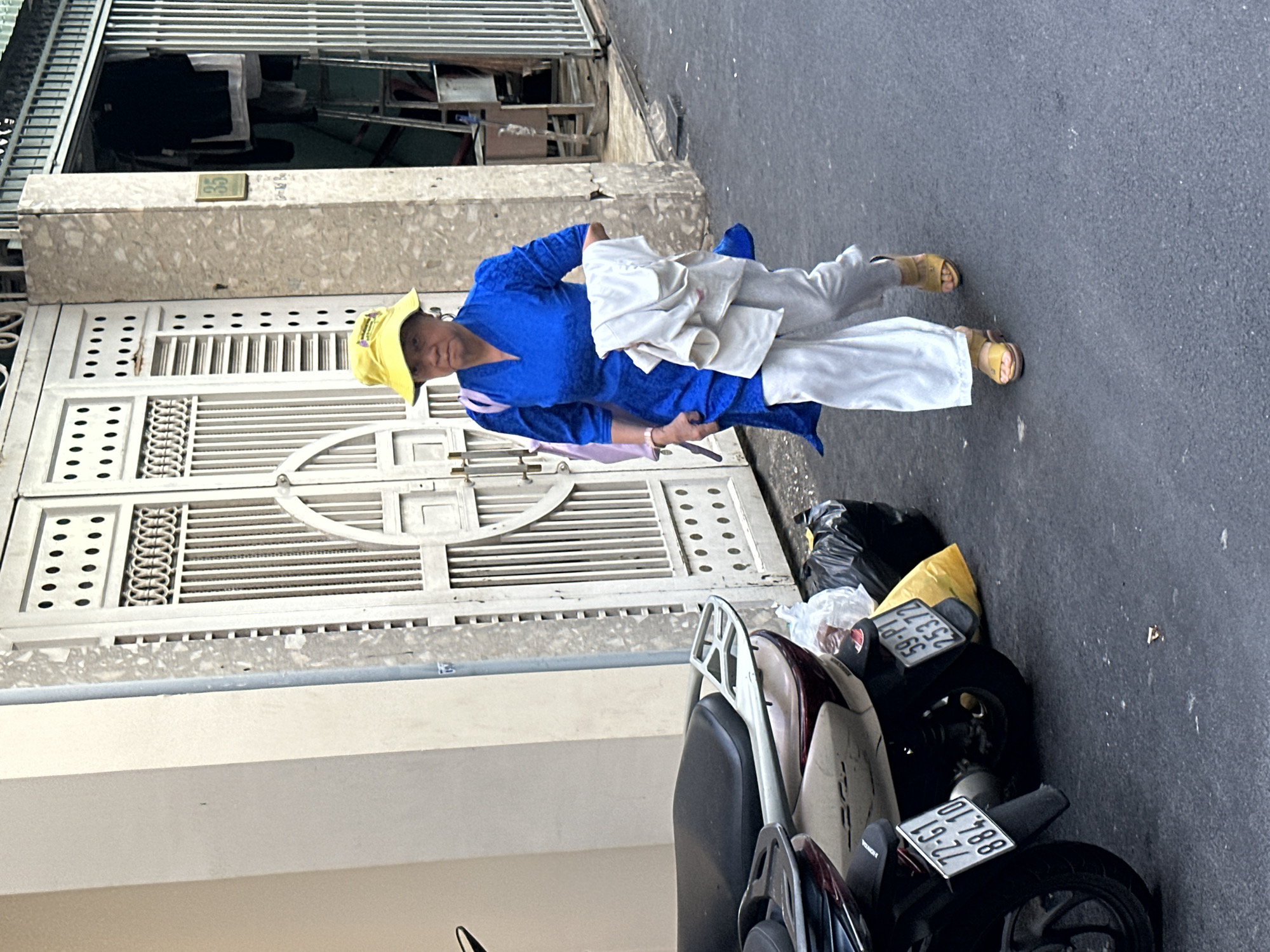 An elderly Asian woman wearing a bright blue tunic, white pants, a yellow hat, and sandals stands on a street in Thủ Đức, Vietnam. She holds a pair of scissors in her right hand and a light-colored garment over her left arm. Behind her is a white metal gate in front of a building, and to her right are two parked motorcycles with black trash bags nearby.