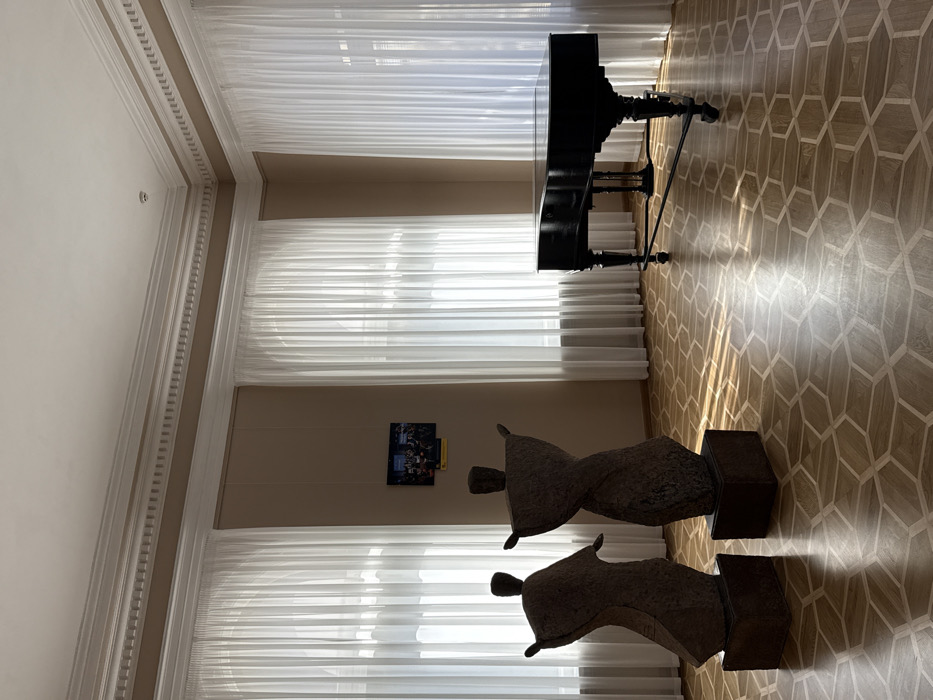 The image depicts an interior view of a room within the National Art Museum of Ukraine in Kyiv. The room features high ceilings and light beige walls adorned with elaborate white crown molding. Three tall, arched windows, draped with sheer white curtains, illuminate the space with natural light, casting distinct patterns on the floor. In the left foreground, two abstract, dark-toned sculptures stand on rectangular pedestals, appearing to be made of coarse material with stylized, raised arms. On the left wall, between two windows, a small digital display screen shows a group of individuals, and a yellow sign is visible beneath it. To the right, a black grand piano with ornate legs is positioned near the windows. The floor is covered in a light-toned wooden parquet, arranged in a geometric pattern of interlocking hexagons and rectangles. No people are present, and the scene is characterized by art objects and musical instruments within a well-lit, architecturally ornate museum setting.