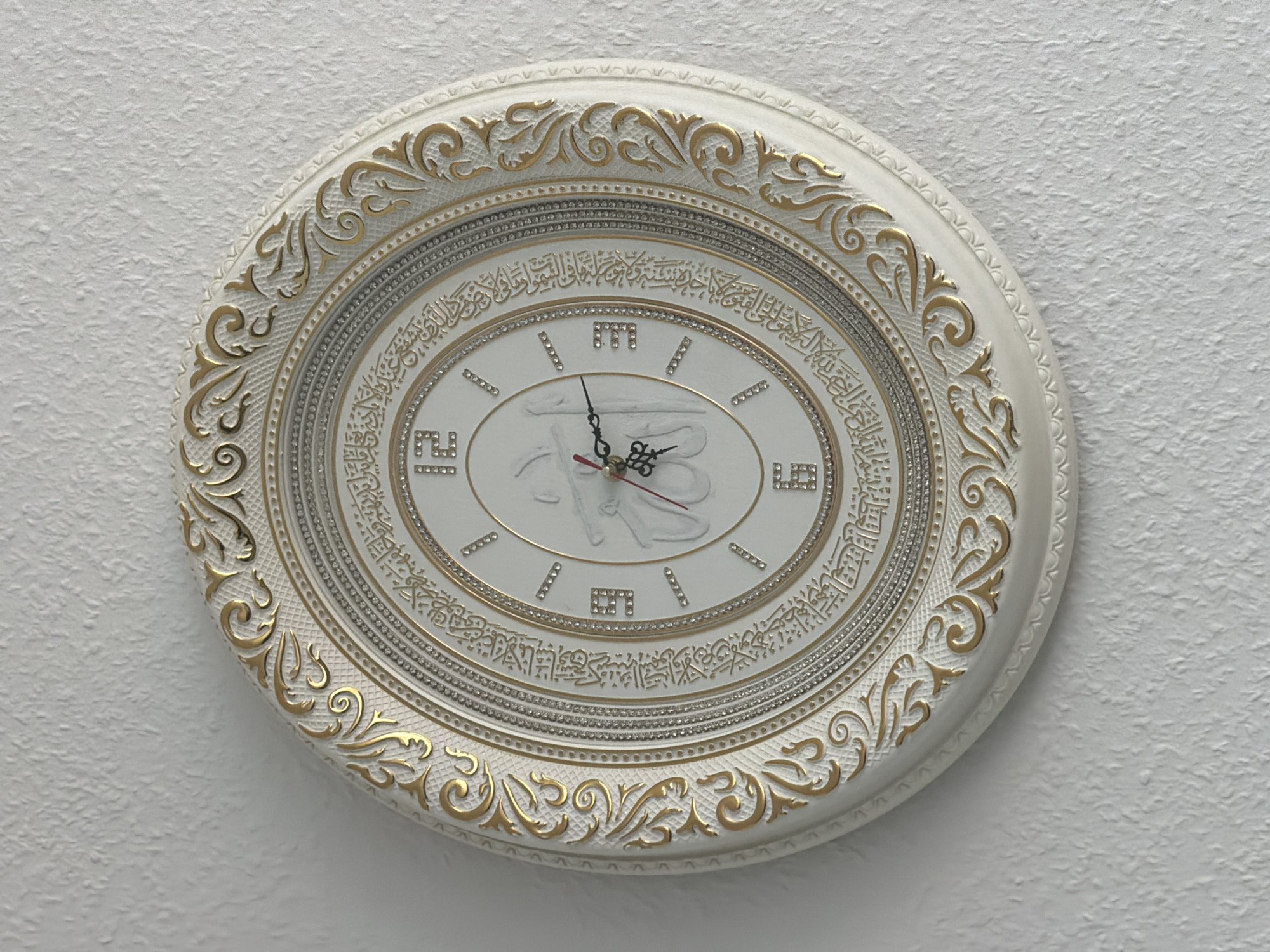 This is a medium shot, eye-level photograph of a decorative wall clock against a textured white wall. The clock has an ornate, oval design with a white frame featuring gold, scroll-like embellishments. The clock face is white and appears to be made of ceramic or a similar material, with Arabic calligraphy encircling the numbers. The numbers themselves are represented by small diamond-like studs and also by stylized Arabic numerals in a square format, with a prominent "9" visible at the bottom left. The clock hands are black and red, and it appears to be showing a time of approximately 10:10. In the center of the clock face, there is a raised, white relief of Arabic script. The lighting suggests it is daytime, likely indoors, given the consistent light on the wall and clock. The setting is a wall in what appears to be a residential or public space in Dorsten, Germany. There are no people visible, and no specific weather or emotional cues are present. The prominent text is in Arabic calligraphy around the clock face and in the central relief.