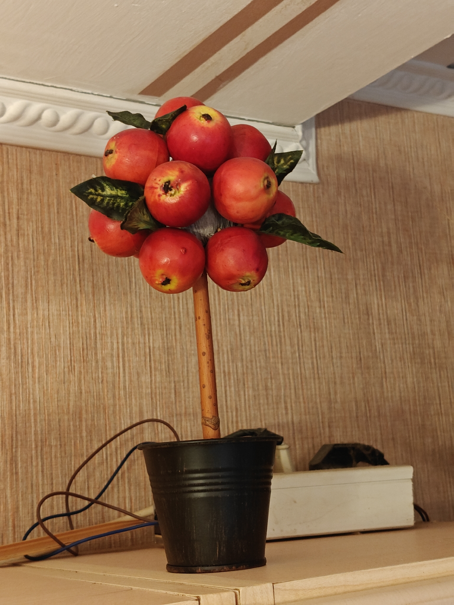 A tabletop decorative item resembling a small apple tree stands in a black pot. The tree has a wooden stick as a trunk, adorned with several red artificial apples and a few green leaves. The pot is placed on a light-colored shelf. In the background, a wall with a wood-grain pattern is visible, and on the shelf behind the pot, a white power strip with a black plug is present. Above the wall, a white ceiling with decorative molding and beige stripes is seen. The scene is static and appears to be indoors, with no people present and no actions taking place.