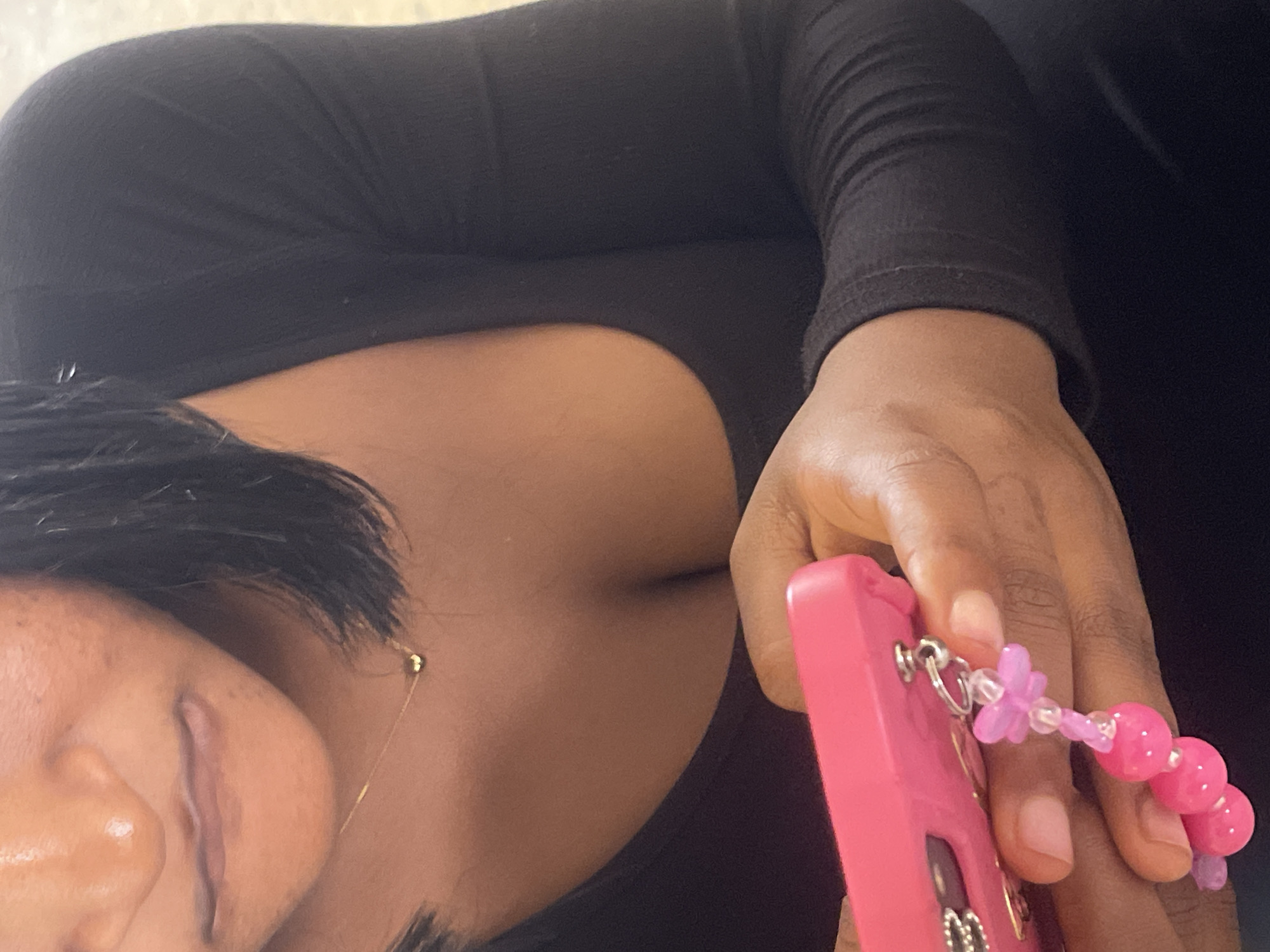 The image shows a close-up of a person holding a pink cell phone with a beaded charm. The person is wearing a black long-sleeved shirt that reveals cleavage. They are also wearing a delicate gold necklace with a small circular pendant. The background is blurred, but appears to be an indoor setting. The lighting suggests the photo was taken during the day. There is no indication of the location being Rafin Par, Nigeria.