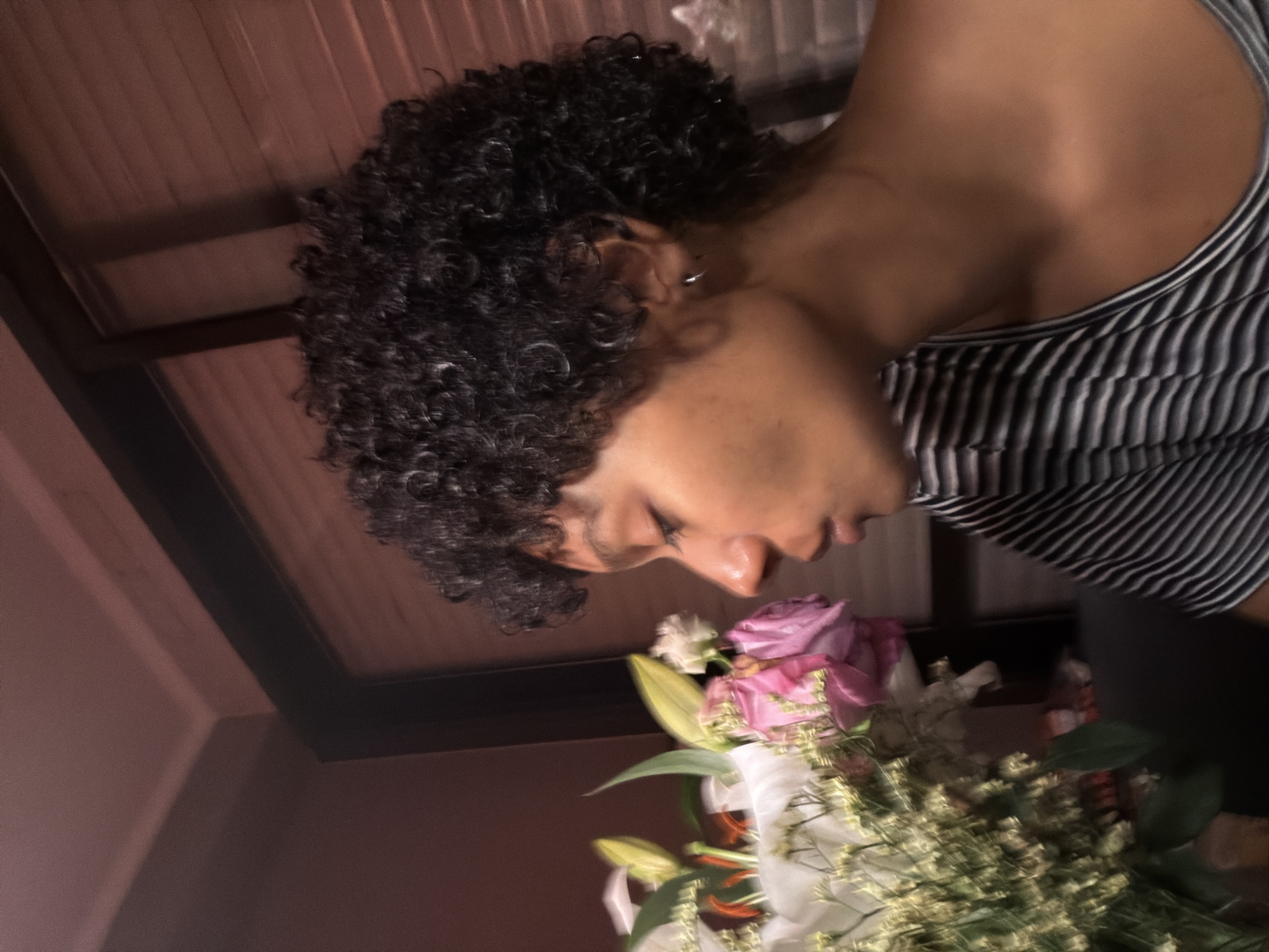 A person with curly black hair is positioned above a bouquet of flowers, which includes roses and lilies. The person is wearing a striped tank top. The background appears to be an interior setting with a partially visible window or ceiling structure. There is no evidence to support the location is in Champs-sur-Marne, France.