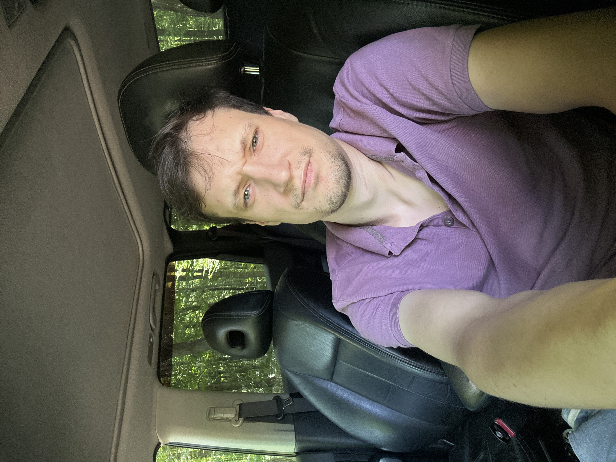 A man with short brown hair and a stubble is sitting in the driver's seat of a car. He is wearing a purple polo shirt. His left arm is extended toward the camera. The interior of the car is visible, including black leather seats, a sunroof, and a seatbelt. Outside the car, trees and foliage suggest a wooded area. The image appears to be a selfie taken in a car, likely parked in a rural or natural setting. The location of Shatava, Ukraine is not depicted or suggested by the visual elements in the image.