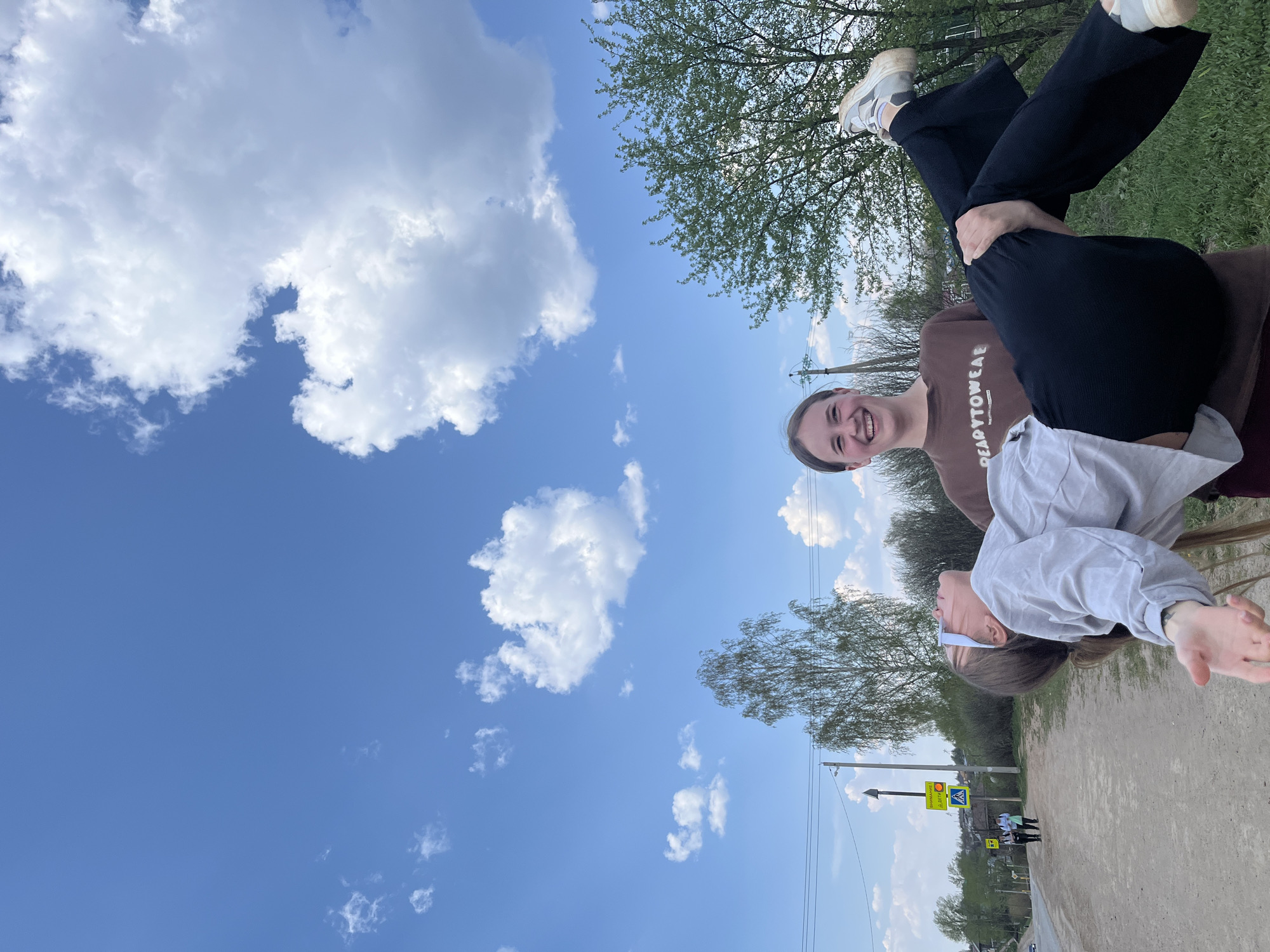 Two women are outdoors on a sunny day in what appears to be a park or recreational area. The location is likely Фоки, Russia. One woman is standing, wearing a brown shirt with white lettering that says "READYTOWEAE" and black pants. The other woman is positioned upside down, supported by the standing woman's shoulders. She is wearing a light gray shirt and black pants with white sneakers. In the background, there is a clear blue sky with scattered white clouds, trees, and glimpses of a street with a pedestrian crossing sign. The standing woman is smiling, while the upside-down woman's face is partially obscured.