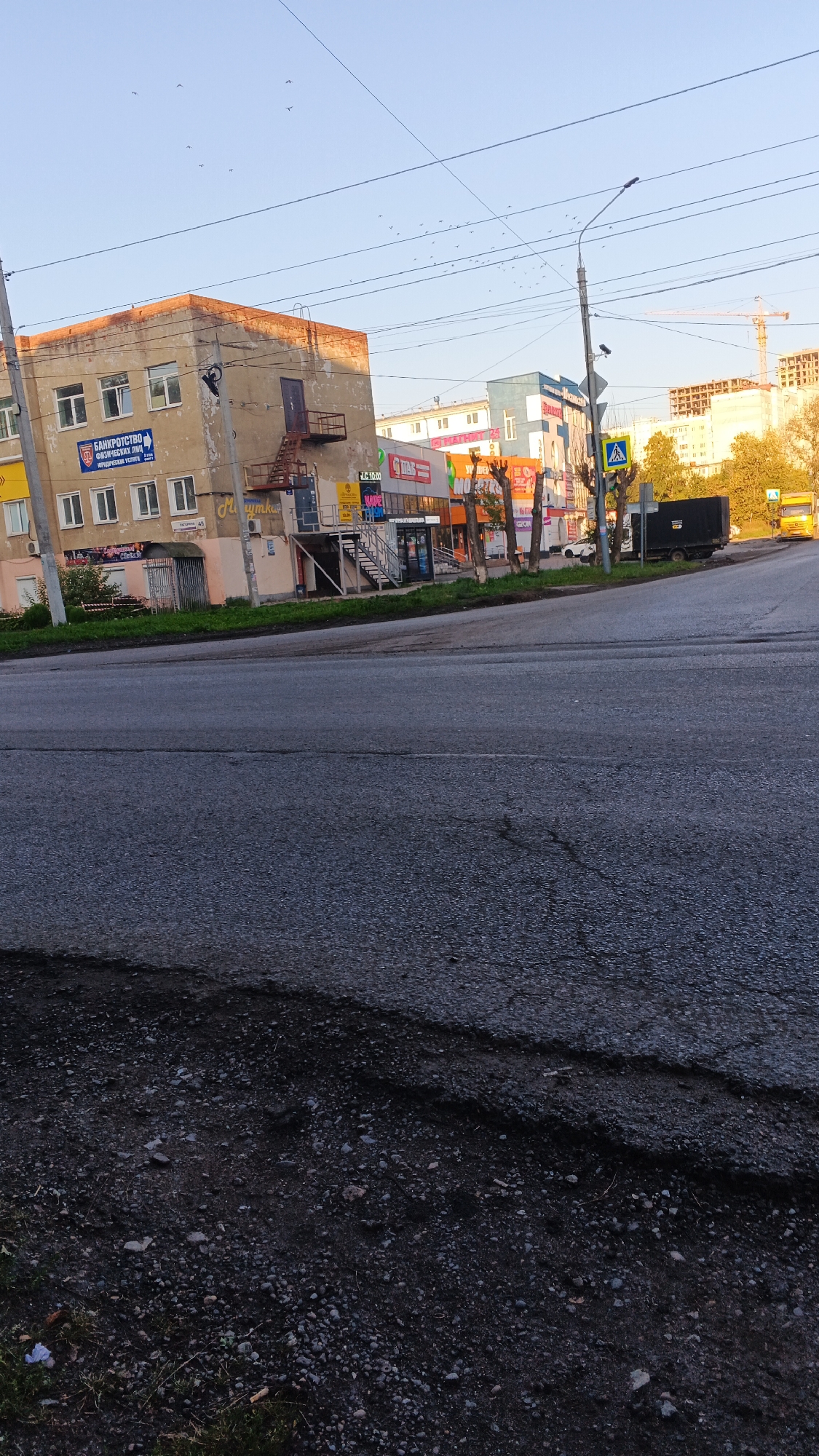 The image depicts a street scene in Izhevsk, Russia, likely in the early morning or late afternoon given the quality of light. In the foreground, a cracked asphalt road dominates the lower half of the frame. Beyond the road, a sidewalk and a grass strip separate the street from a row of buildings.
The buildings are varied in architectural style and condition. The leftmost building is a multi-story structure with a weathered, orange-tinged facade and numerous windows. It appears to have a commercial ground floor with signs. One prominent sign reads "БАНКРОТСТВО ФИЗИЧЕСКИХ ЛИЦ" (Bankruptcy for individuals). Further along the street, other buildings feature more modern facades with prominent signage for businesses such as "МАТРИЦА 24" and what appears to be a cafe or restaurant.
Several vehicles are visible. A dark-colored truck is parked on the right side of the road, near a pedestrian crossing sign. Further down the street, a yellow truck is driving away from the camera. A white car is also parked or moving on the road.
The sky is clear and pale blue, with a few scattered birds visible. Overhead, power lines and streetlights are strung across the scene, indicating an urban environment. In the background, a construction crane is visible, suggesting ongoing development. The overall impression is a typical urban street with a mix of older and newer structures, commercial activity, and infrastructure.