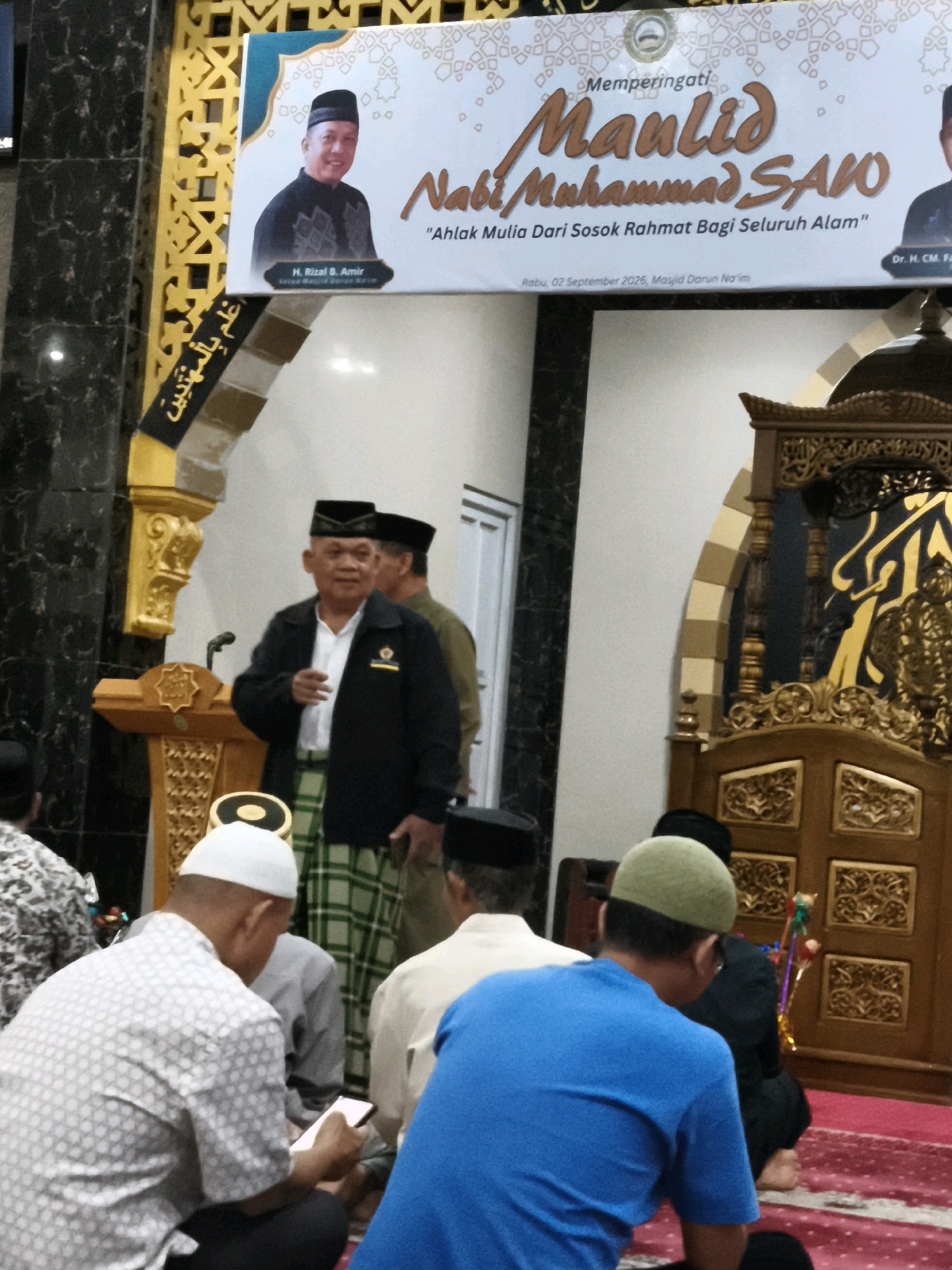 This image depicts an indoor event, likely a religious gathering, within a building that appears to be a mosque or community hall, potentially located in Makassar, Indonesia. In the foreground, several men are seated on a red carpet, facing away from the camera. One man in a white patterned shirt is looking at a mobile phone. Another man is wearing a blue t-shirt.
In the mid-ground, a man in a black jacket, white shirt, and a checkered sarong is standing behind a wooden podium, addressing the attendees. He is wearing a black cap. Behind him, another man in a similar black cap and a greenish-brown uniform is partially visible.
The background features a large, ornately decorated banner with text in Indonesian. The banner includes images of two men, one in the foreground of the banner and another to the right. The text on the banner appears to be related to a Maulid (Prophet Muhammad's birthday) celebration. To the right of the podium, there is a section of a wooden structure with intricate carvings, possibly part of the mosque's interior or a decorative element. The overall lighting suggests an indoor setting with artificial illumination.