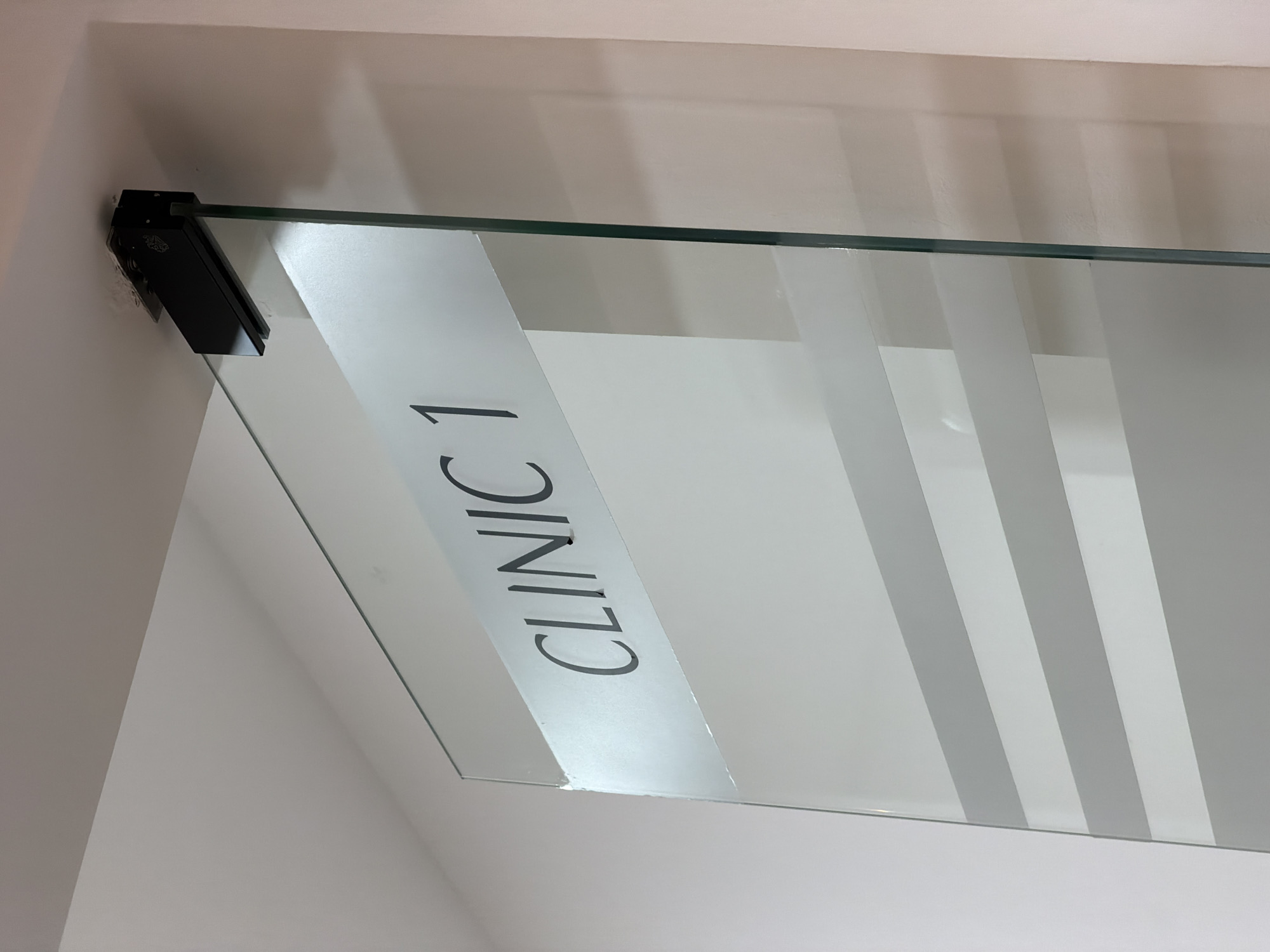 The image shows a close-up view of a glass door with the text "CLINIC 1" frosted onto it. The door is suspended from a black metal fixture attached to a white ceiling. The glass has horizontal frosted stripes applied to it. The background is a plain white wall with some subtle shadows. There are no people or other objects clearly visible. The location is Sharm El Sheikh, Egypt.