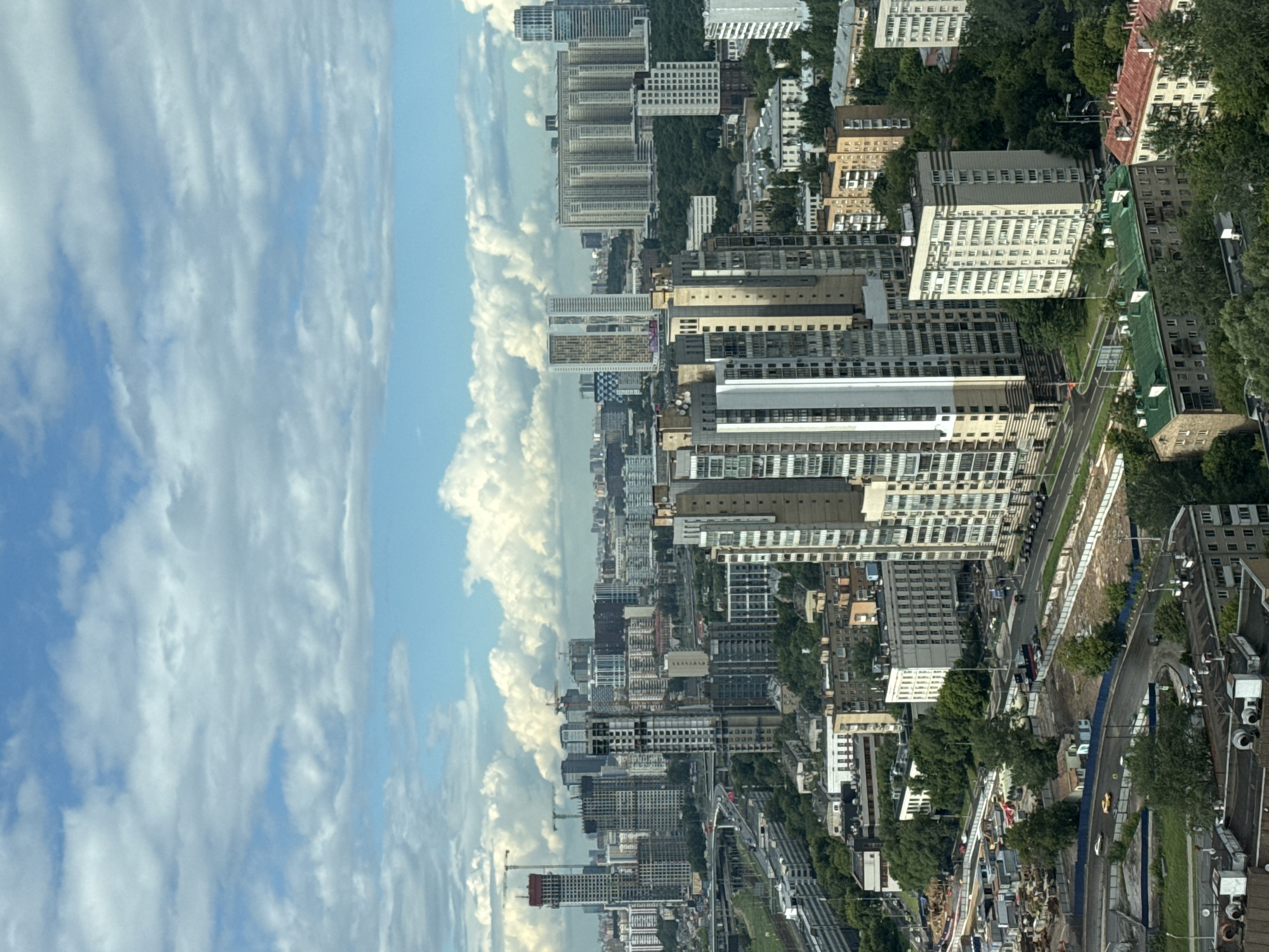 This is a vertical, high-angle aerial shot of a cityscape in Khimki, Russia. The scene is dominated by a dense collection of apartment buildings, varying in height and architectural style, interspersed with patches of green trees. The buildings are a mix of modern high-rises with many windows and older, more utilitarian structures. Some buildings appear to be under construction, indicated by cranes and scaffolding.
A road or highway runs through the middle of the scene, with some vehicles visible, including what appears to be a yellow car. There are also indications of infrastructure like power lines and possibly train tracks. The overall impression is a developing urban environment with a blend of residential and commercial structures.
The sky on the left side of the image is filled with dramatic, swirling clouds, suggesting a dynamic weather pattern. The lighting indicates it is daytime, likely mid-morning or afternoon, given the brightness and the shadows cast by the buildings and trees. There is no visible text in the image. The scene conveys a sense of urban scale and activity, with the vastness of the sky contrasting with the man-made structures.