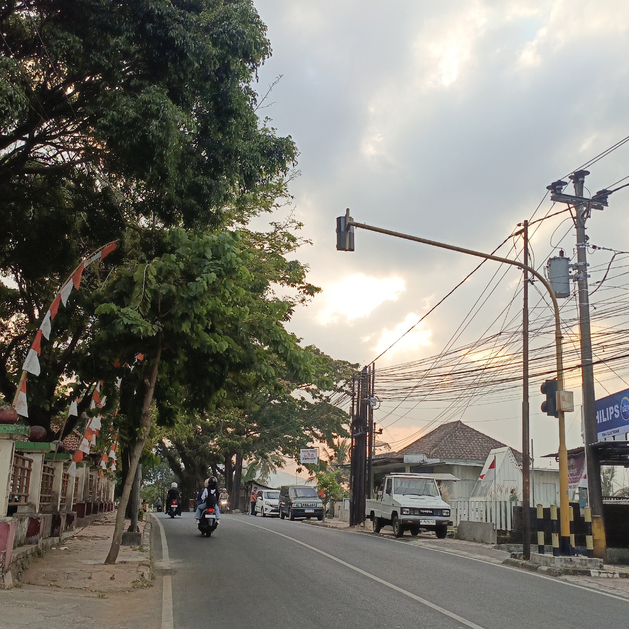 This is a street-level, eye-level medium shot of a road in Salatiga, Indonesia, during the day. The scene captures a moderately busy street with vehicles and pedestrians. On the left side of the road, there's a sidewalk with a low, decorative wall. Red and white Indonesian flags are strung along the top of this wall. Lush green trees line the sidewalk, their branches extending over the road and sidewalk, partially obscuring the sky.
The road itself is paved and has white lane markings. Several motorcycles are present, with two riders visible in the foreground. One motorcycle is in the middle lane, and another is further down the road. A white Toyota pickup truck is parked on the right side of the road. Behind the truck, a black SUV is also parked. Further down the street, there are buildings and more trees.
Electrical poles with numerous wires crisscross the upper part of the image, contributing to a dense urban feel. A traffic light is visible, suspended over the road. The sky is cloudy, with the sun attempting to break through, casting a warm, diffused light on the scene, suggesting it might be late afternoon. A sign for "BAKSO JUMBO" is visible on a building in the background. The overall impression is one of everyday life unfolding on a typical Indonesian street.