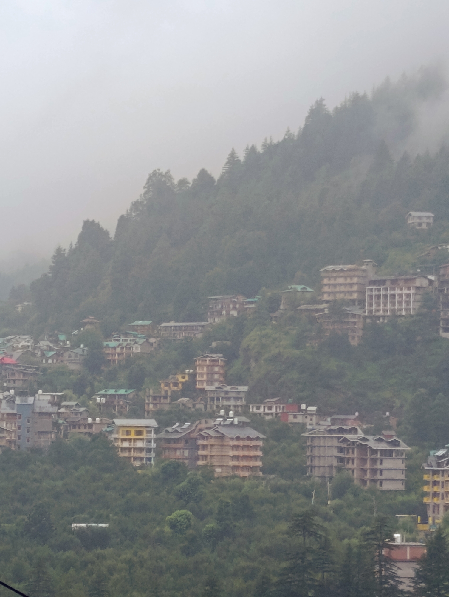 A hazy, overcast day looms over a hillside settlement in Vashisht, India. The scene is dominated by numerous buildings, varying in height and architectural style, nestled amongst lush green trees and foliage that covers the steep terrain. The buildings appear to be multi-storied structures, some displaying balconies and visible windows, while others are under construction, their concrete frameworks exposed. A few buildings are painted in muted tones of beige and yellow, while others have grey or green roofs. The overall impression is of a densely populated residential or hotel area built on a challenging, sloped landscape. The fog and mist in the background obscure the full extent of the settlement and the surrounding mountains, giving the scene a somewhat muted and subdued atmosphere. There are no people or vehicles clearly visible in the image.