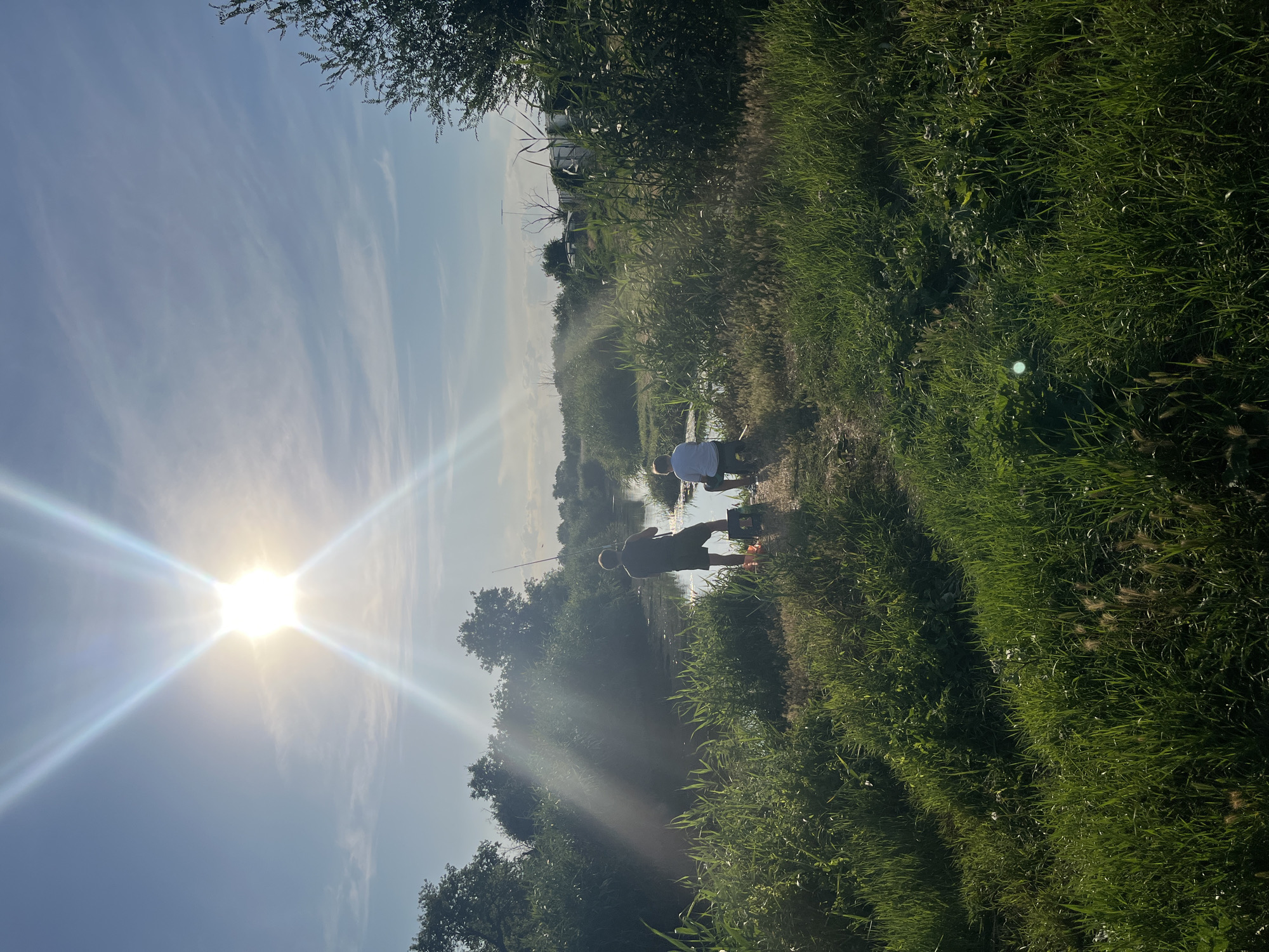 The image shows two figures fishing in a narrow body of water, possibly a river or canal, in a rural or semi-rural setting. The sun is bright and positioned in the upper left quadrant of the image, creating lens flare and casting strong light. The foreground is dominated by tall, green grass and reeds, suggesting a natural environment. In the mid-ground, a person wearing a dark t-shirt and shorts stands casting a fishing rod, with a bucket at their feet. To their right, a second person, wearing a light-colored t-shirt and shorts, is seated, also fishing. The water is calm and reflects the sky and surrounding vegetation. The background consists of dense trees and shrubs on either side of the water, with a few structures, including what appear to be outbuildings or temporary structures, visible in the distance on the right. The sky is blue with wispy clouds. The scene depicts a peaceful outdoor activity, likely during daytime. The location in Яново-Грушевский, Russia, is inferred from the provided context, but the visual elements themselves are generic to many temperate rural landscapes.