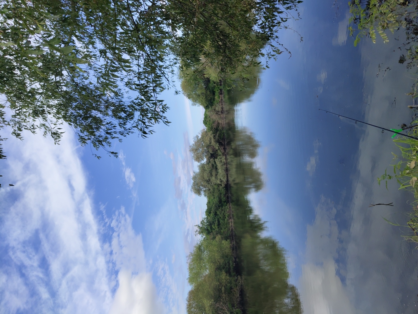 The image depicts a serene natural landscape with a calm body of water reflecting the sky and surrounding trees. The sky is a vibrant blue with scattered white clouds, creating a picturesque scene. On the left side of the image, lush green trees and bushes line the bank of the water. The water itself is still and dark, with clear reflections of the sky and foliage. In the foreground on the right, a fishing rod is set up, indicating recreational activity. The environment suggests a peaceful outdoor setting, possibly a lake or a slow-moving river, located in Krushare, Bulgaria. The overall atmosphere is tranquil and natural.