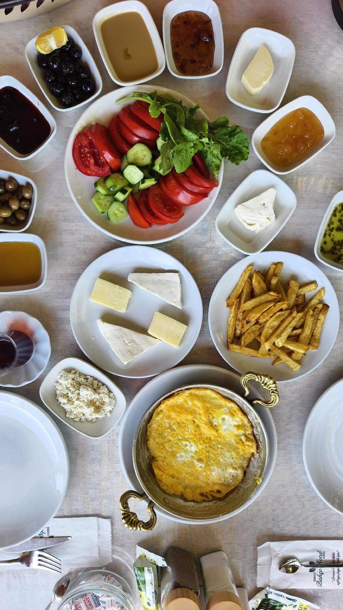 A Turkish breakfast spread is laid out on a table covered with a textured, light-colored cloth, likely in a cafe or restaurant in Selçuk, Turkey, known for its historical significance and culinary traditions. The overhead shot reveals a variety of dishes arranged across the frame.
In the upper left quadrant, a small white dish contains black olives, one halved lemon slice, and a sprig of green herb. Adjacent to it, a rectangular white dish holds a smooth, tan spread, possibly tahini or a similar paste. To its right, another rectangular dish contains a dark, jam-like substance with visible fruit pieces.
A large white oval plate in the center is piled with sliced ripe tomatoes, cucumber slices, and fresh green leaves, likely arugula or parsley, creating a vibrant salad. Below this, on a smaller white plate, are several pieces of cheese: two triangular white pieces and two rectangular, pale yellow pieces.
To the right of the salad plate, a small white dish holds a light orange jam. Below that, another white rectangular dish contains a cube of white cheese. Further to the right, a deep white dish holds a vibrant green mixture, possibly a herb salad or olive oil with herbs.
A small glass of dark red liquid, likely Turkish tea, sits on a saucer to the lower left of the salad plate. Below the tea, a small white dish is filled with crumbled white cheese, resembling feta.
A prominent plate in the lower half of the image holds a generous portion of golden-brown french fries. Below the fries, a round metal pan with decorative handles contains a fried egg, cooked sunny-side up with a slightly browned white and a runny yolk.
Other elements on the table include white plates, cutlery, salt and pepper shakers, and packets of sugar and napkins. The overall impression is a bountiful and traditional Turkish breakfast, highlighting fresh ingredients and diverse flavors.