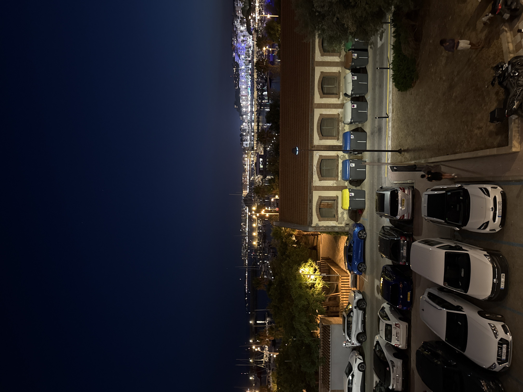 An aerial shot captured at night in the city of Blanes, Spain, shows a street with parked cars and a waterfront view. The sky is a deep, dark blue, with a distant city skyline illuminated by numerous lights. In the foreground, a row of various types of cars are parked. Among them are a white Audi sedan with license plate 1040 CZC, a white van with license plate 4335 JHM, and a white Toyota Yaris with license plate 6079 LXD. Further up the street, there are more cars, including a blue hatchback and several white SUVs. Adjacent to the parking area, a row of large, multi-colored bins (yellow, blue, grey, and brown) are visible, situated in front of a building with distinctive arched windows. Behind the building, trees are illuminated by streetlights, and beyond that, a harbor filled with sailboats stretches towards the illuminated city. Two individuals are present: one person in dark clothing walks along the sidewalk, while another person in a light-colored shirt and shorts stands near the bins. A motorcycle is parked in the lower right corner of the image.