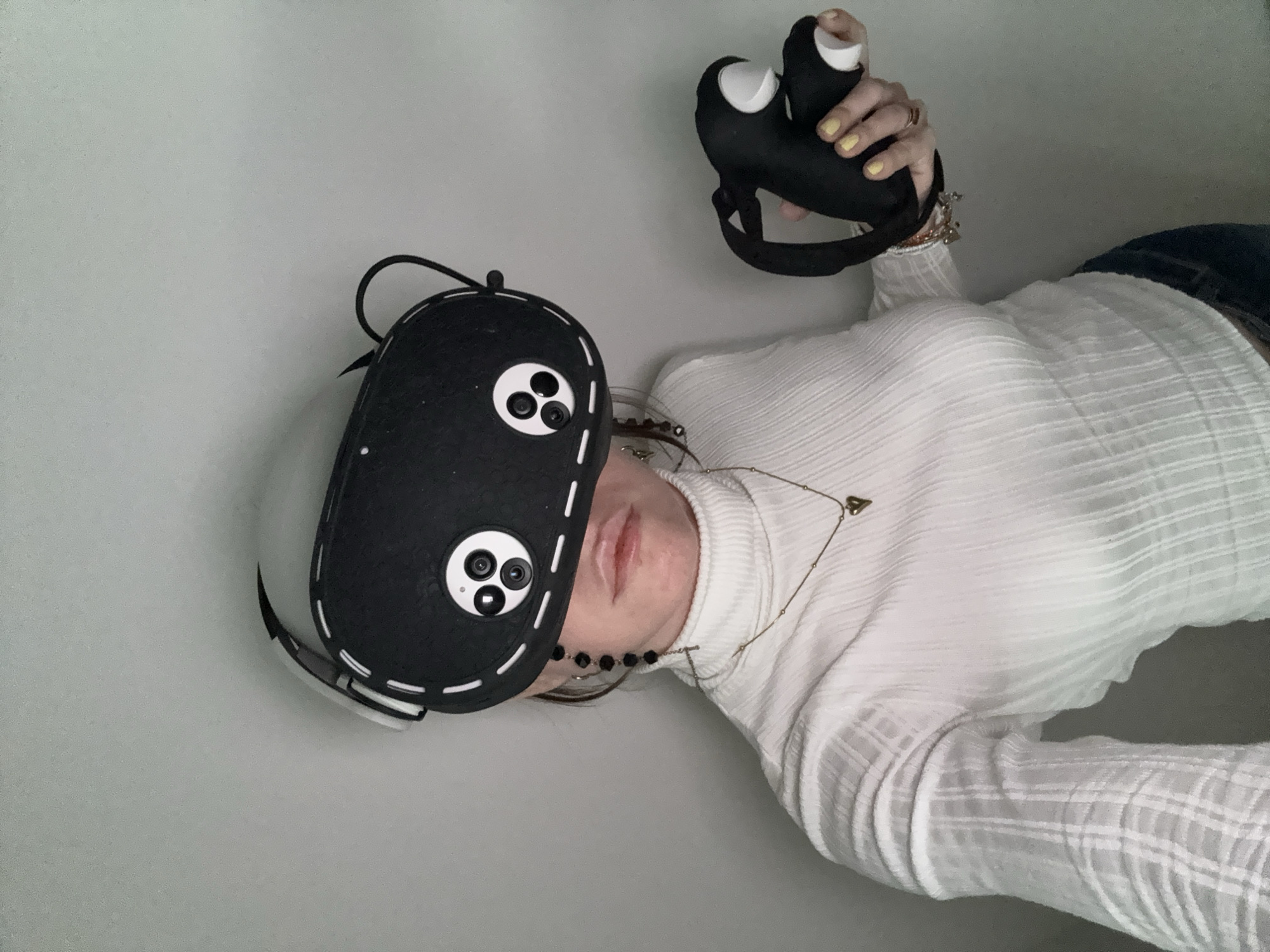A woman wearing a white turtleneck sweater is lying down on a gray surface. She is holding a black virtual reality (VR) headset in her right hand and another VR headset is partially obscuring her face. The headset features a black cover with faux camera lenses. She is wearing a gold necklace and several bracelets. Her fingernails are painted yellow. The background is a plain, light gray surface. There is no visible indication of the image's location being Moscow, Russia.