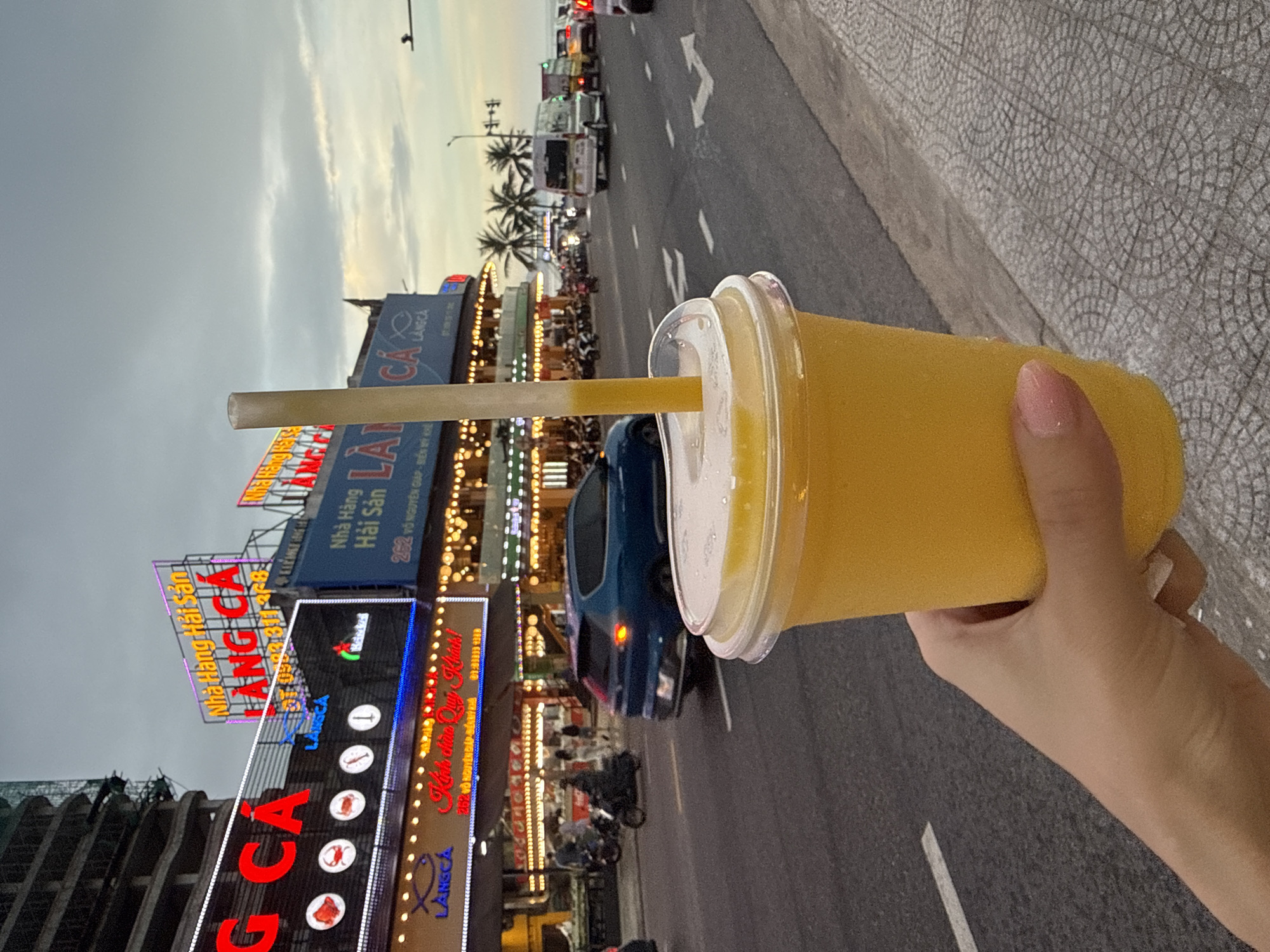 An upright outdoor shot depicts a hand holding a clear plastic cup filled with a yellow beverage and a straw, against a cityscape background. The cup is prominently positioned on the right side of the frame.
In the background, a road with light vehicular traffic runs parallel to the street with palm trees lining the roadside. On the left side, lit signs indicate various commercial establishments. Specifically, signs such as "LANGCA" and "Nhà Hàng Hải Sản" are visible, along with decorative lighting. The skyline above the cityscape suggests twilight or dusk. The foreground shows a sidewalk with a textured surface.