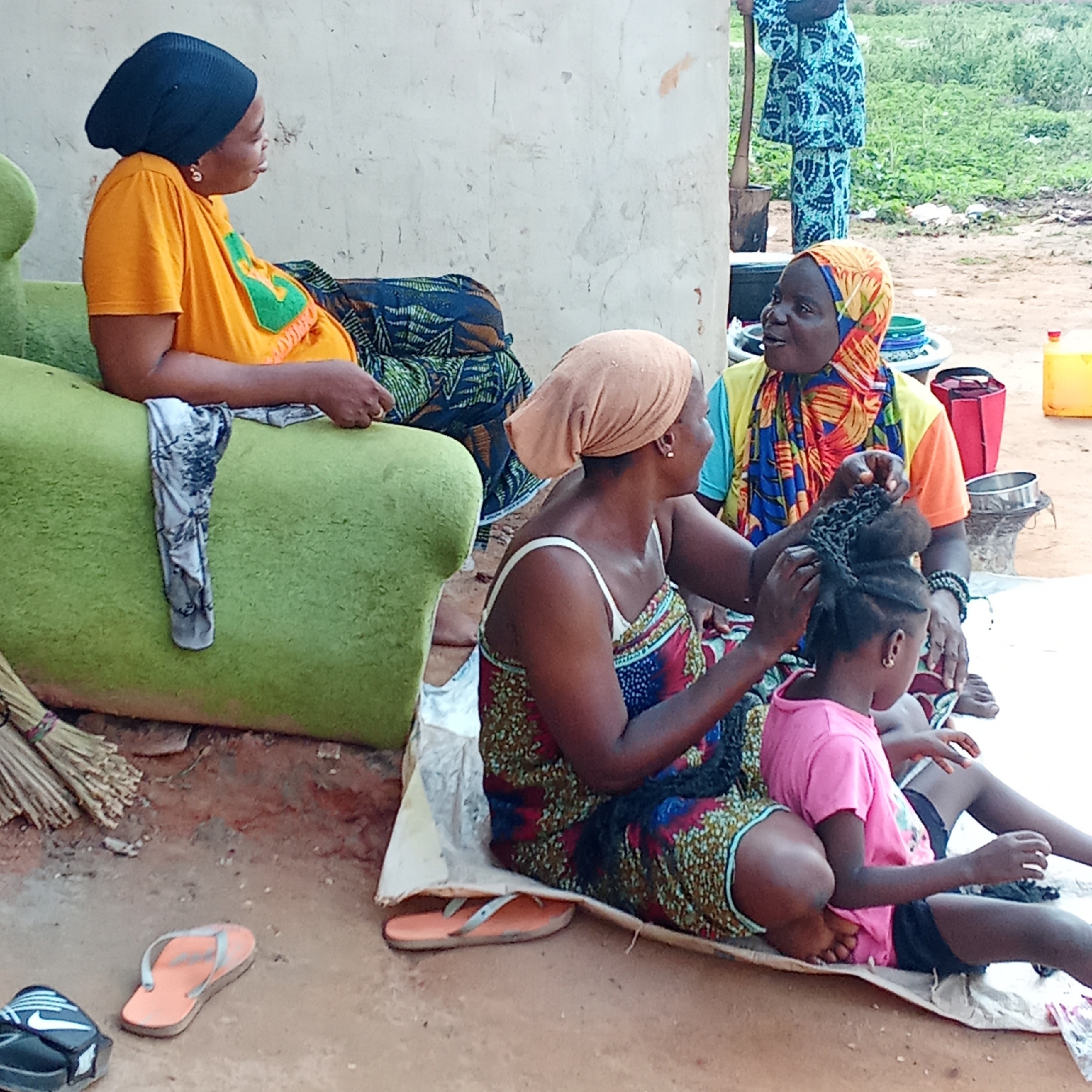 In Doma, Nigeria, the photo shows four women and a child. One woman in an orange t-shirt and black head covering sits on a green couch, talking to another woman with a colorful headscarf and orange shirt who is styling a young girl's hair. The girl wears a pink shirt. A third woman in a patterned dress assists with the hair styling. Another woman stands in the background, partially obscured. Various household items are scattered around the scene, including flip-flops, a broom, pots, and containers. The setting appears to be outdoors, adjacent to a building.