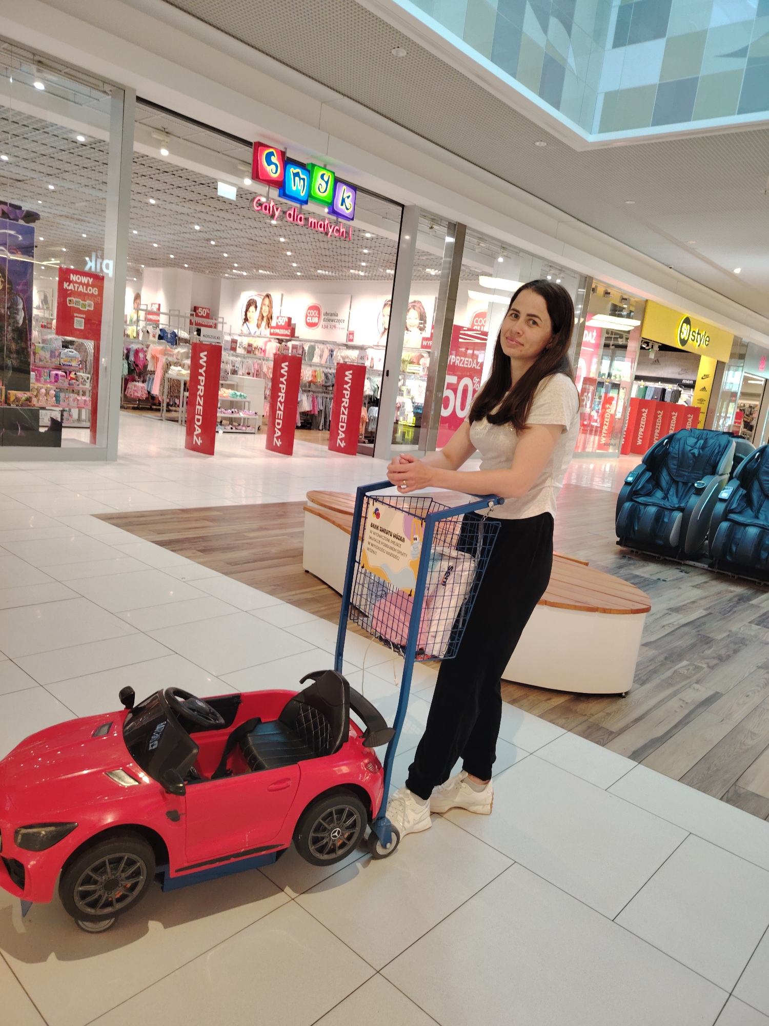 A woman stands in a shopping mall in Wołomin, Poland, holding the handle of a shopping cart. In front of the cart is a small, red toy car, likely a child's ride-on vehicle. The woman is wearing a light-colored t-shirt and black pants, with white sneakers. She is looking towards the camera with a slight smile. The shopping cart is blue and has a wire basket filled with items, including a white bag and what appears to be pink fabric. Behind the woman, the mall environment is visible, with store entrances, displays of merchandise, and seating areas. The floor is tiled with a combination of light and wood-patterned tiles. A prominent sign above a store entrance reads "Smyk," with Polish text below. The overall lighting suggests an indoor, well-lit environment.