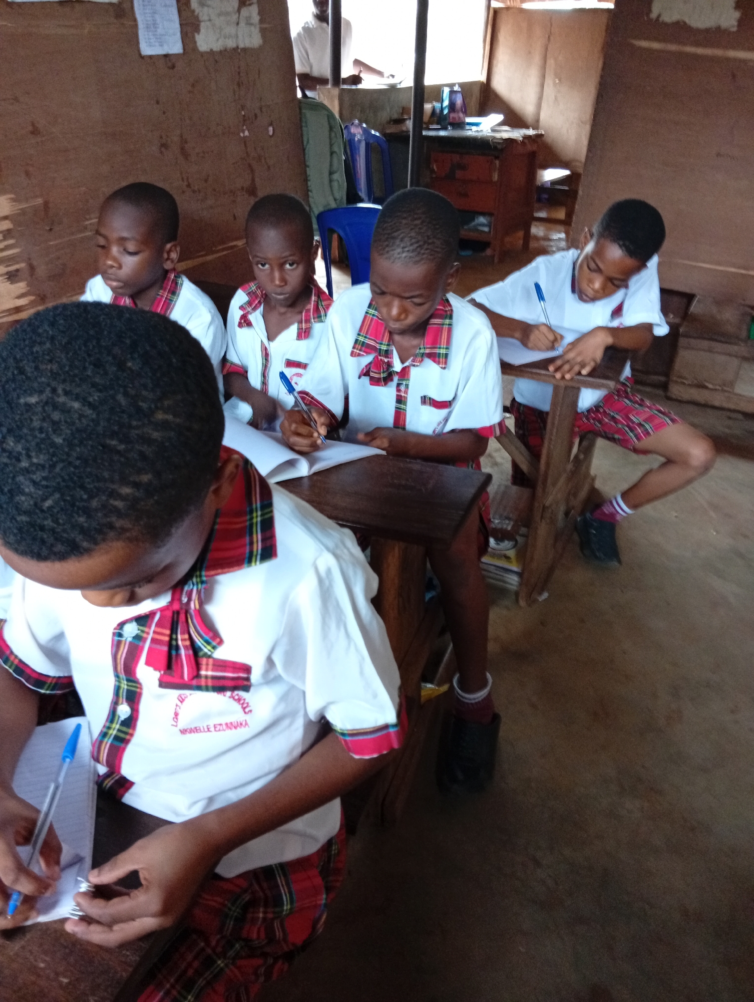 Four children are seated at desks in a classroom in Unknown, Unknown. They are wearing school uniforms with white shirts and red, green, and yellow plaid trim. Three of the children are writing in notebooks with blue pens. The fourth child is looking directly at the camera. A man is standing in the background, partially obscured by a wooden wall. There is a blue plastic chair and a desk in the background as well. The room appears to be sparsely furnished and may be a basic classroom setting.