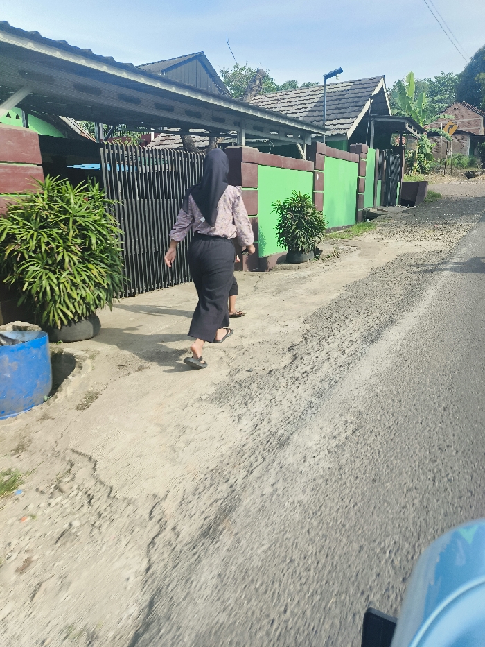 The image shows a woman wearing a black headscarf and a long black skirt walking on a paved road in Jatiluhur, Indonesia (latitude: -6.576, longitude: 107.438). The woman is walking away from the camera and is followed by a young girl. The woman is wearing a light colored shirt and black pants and the girl is wearing a light colored shirt and dark pants. The road is lined with a low concrete wall with a green painted surface and there are several trees and plants growing near the wall. The woman and girl are walking on the right side of the road and there is a blue vehicle parked on the left side of the road, only a part of the vehicle is visible. The image is captured from a moving vehicle. The scene is daytime and the sky is blue. The road is slightly uneven with some areas of cracked asphalt and some areas of dirt. The woman and the girl are the only people visible in the image.